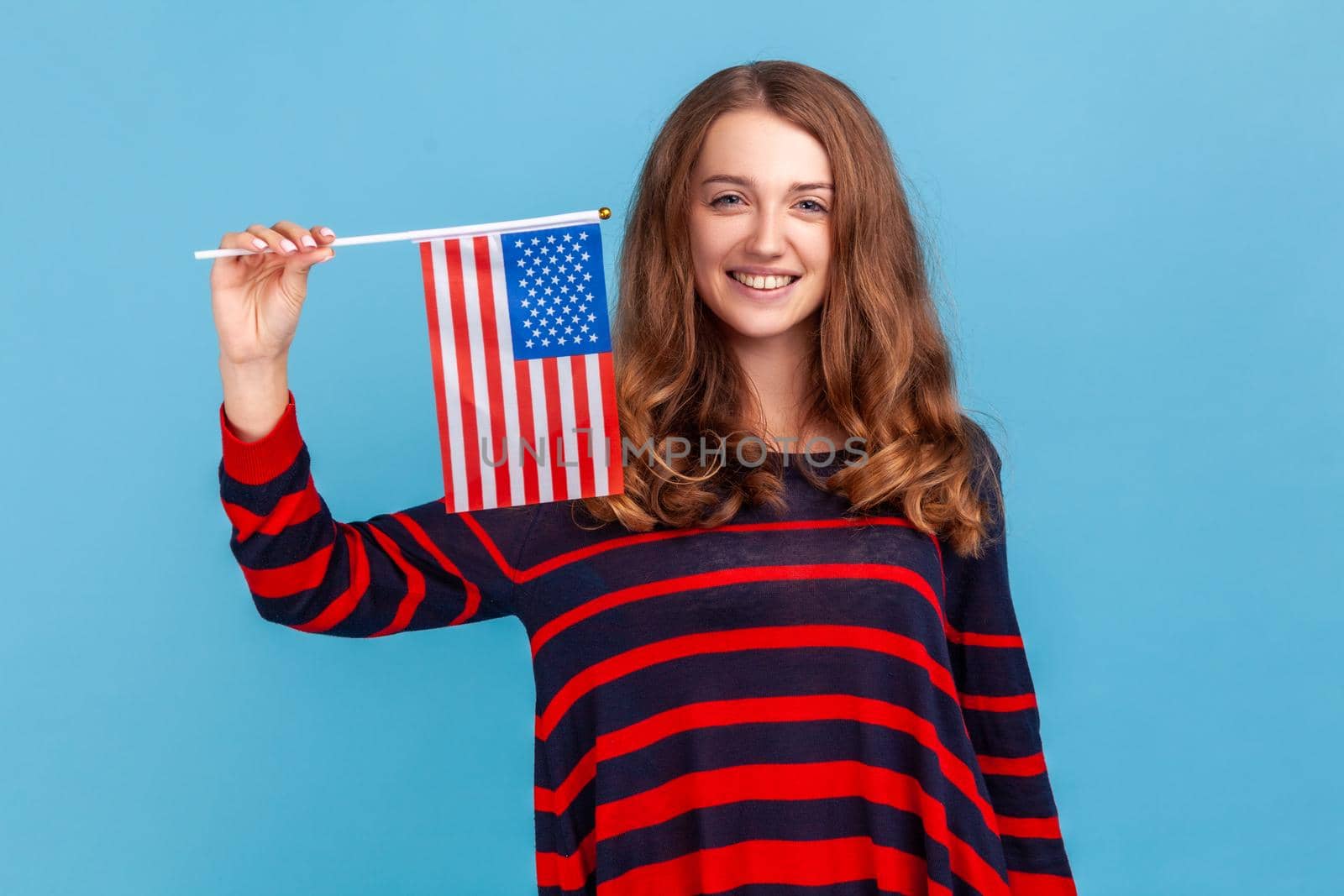 Good looking woman wearing striped casual style sweater, waving united states of america flag, celebrating national holiday, patriotism, independence. Indoor studio shot isolated on blue background.