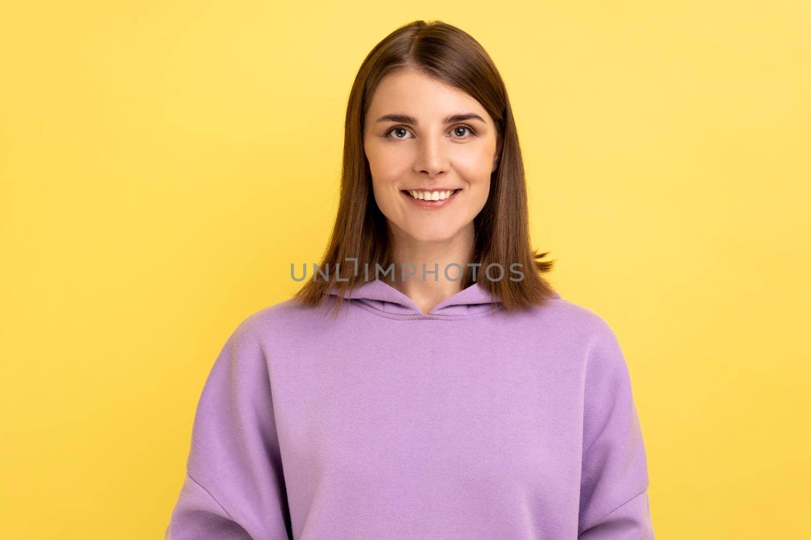 Portrait of happy successful woman standing, looking at camera with charming and joyous toothy smile, expressing happiness, wearing purple hoodie. Indoor studio shot isolated on yellow background.