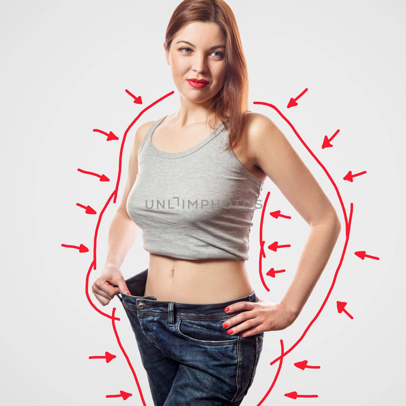 portrait of happy beautiful slim young woman in big jeans and gray top showing successful weight loss, indoor, studio shot, isolated on light gray background, diet concept.