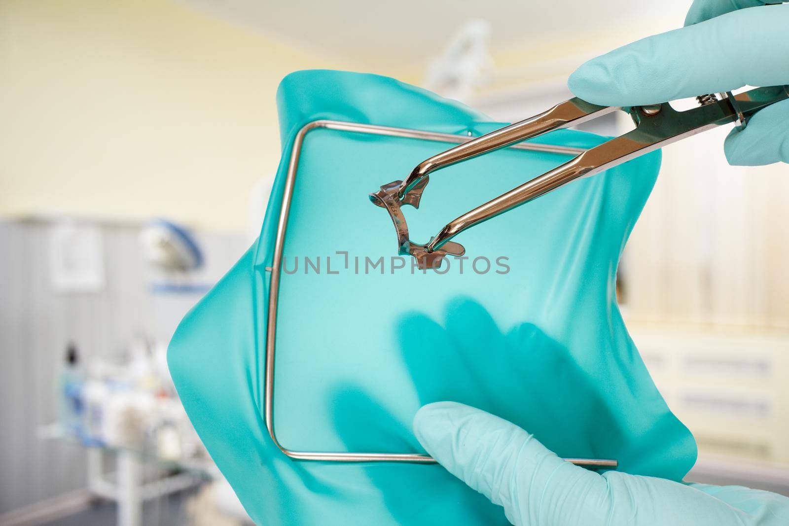 Dentist's hands in latex gloves with a rubber dam clamp forceps, a rubber dam and a metal frame with a detal clinic on the background. Medical tools concept.