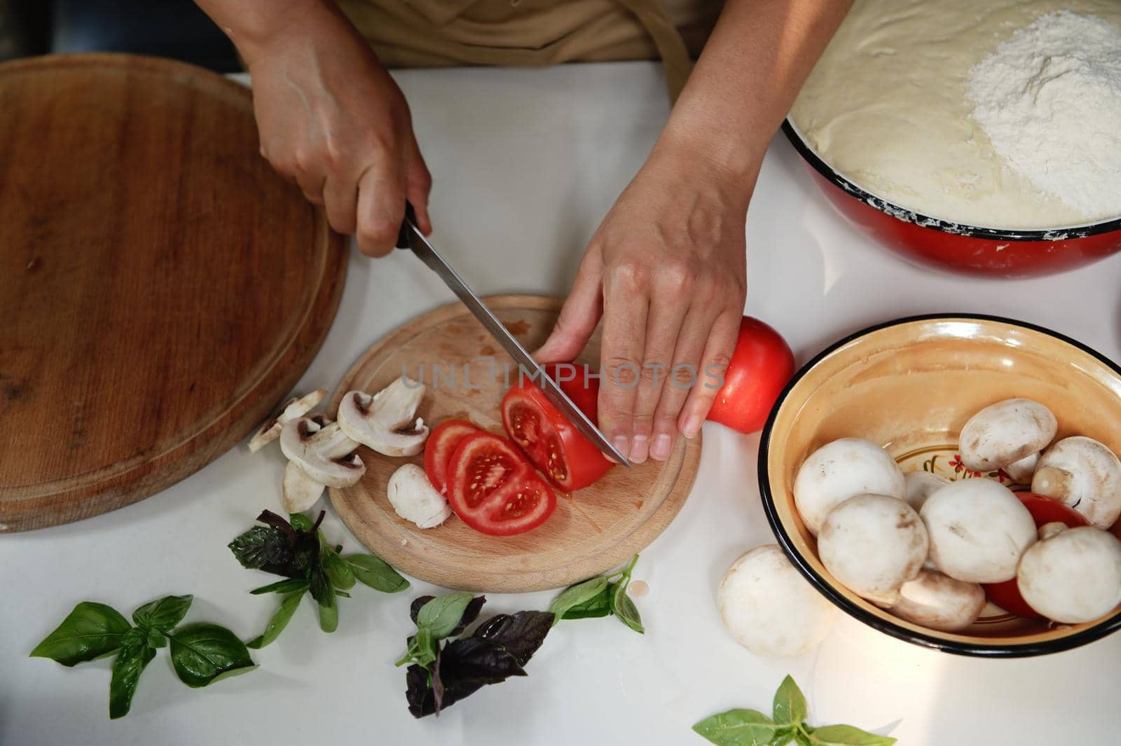 Top view of chef's hands slicing fresh ripe tomatoes with kitchen knife on cutting board, next to chopped mushrooms and basil leaves on table while preparing vegetarian healthy food in rustic kitchen