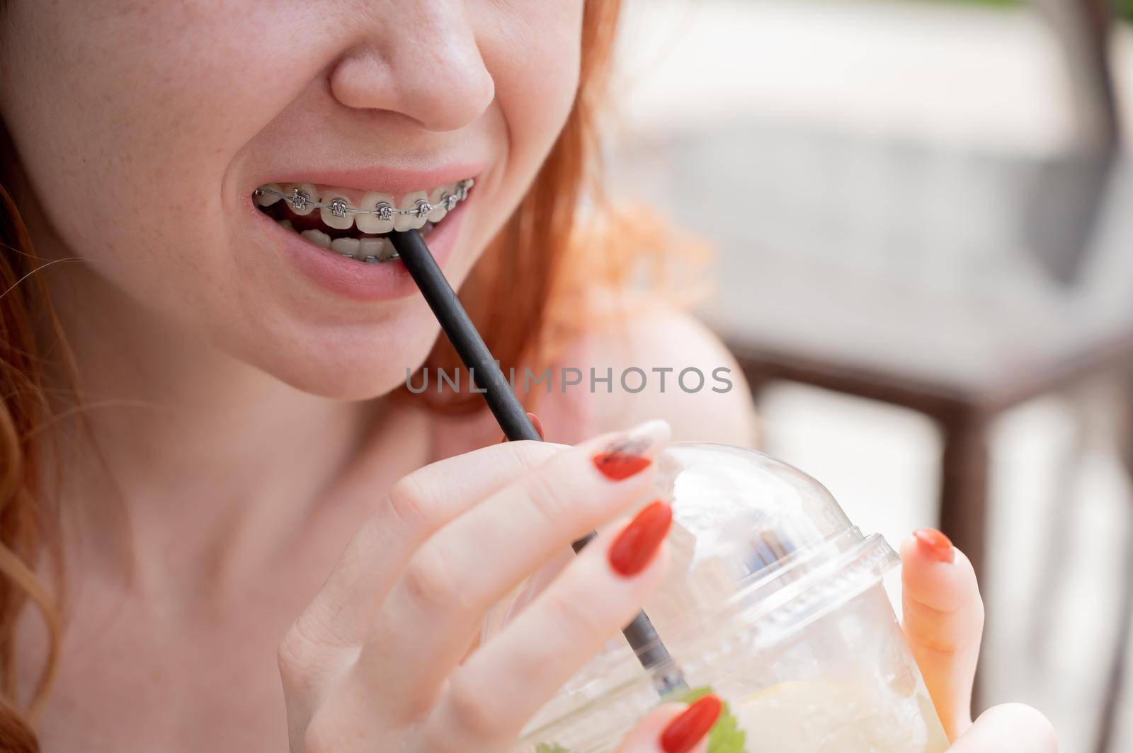 Young beautiful red-haired woman with braces drinks cooling lemonade outdoors in summer. Portrait of a smiling girl with freckles