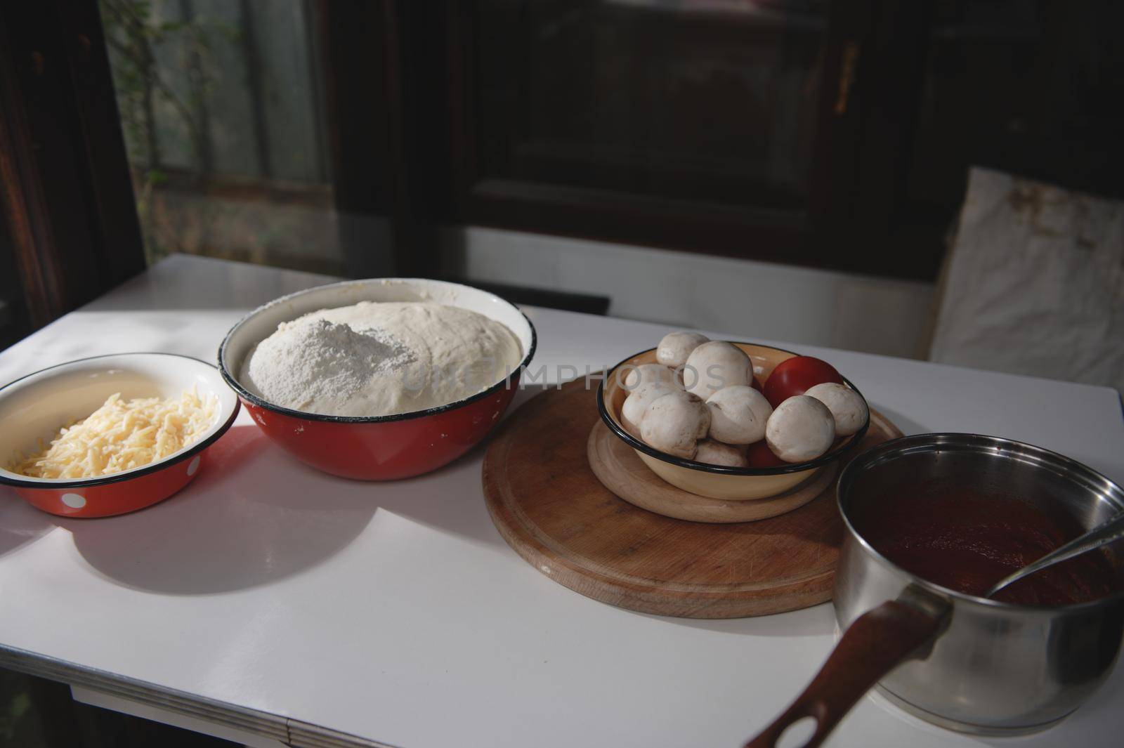 Assortment of cooking ingredients for preparing homemade pizza : rising yeast dough, grated cheese, fresh champignons and tomatoes in enamel bowls on the table in rustic kitchen. Food still life