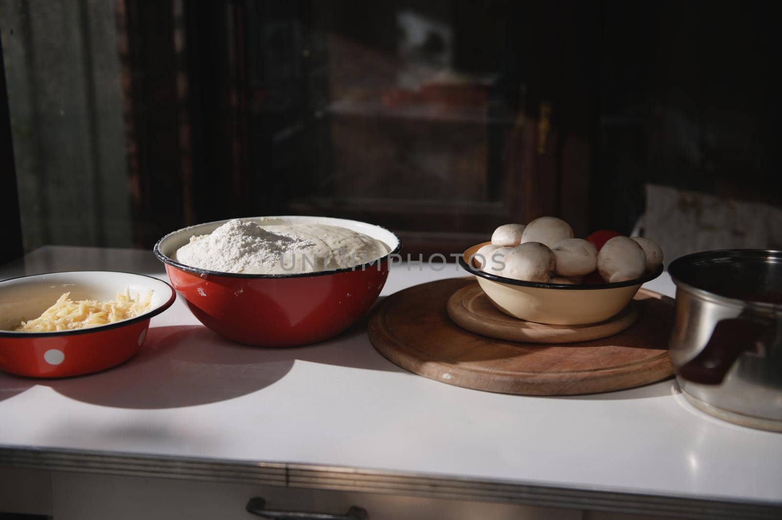 Assortment of baking ingredients for preparing homemade pizza : rising yeast dough, grated cheese, fresh champignons and tomatoes in enamel bowls on the table in rustic kitchen. Food still life