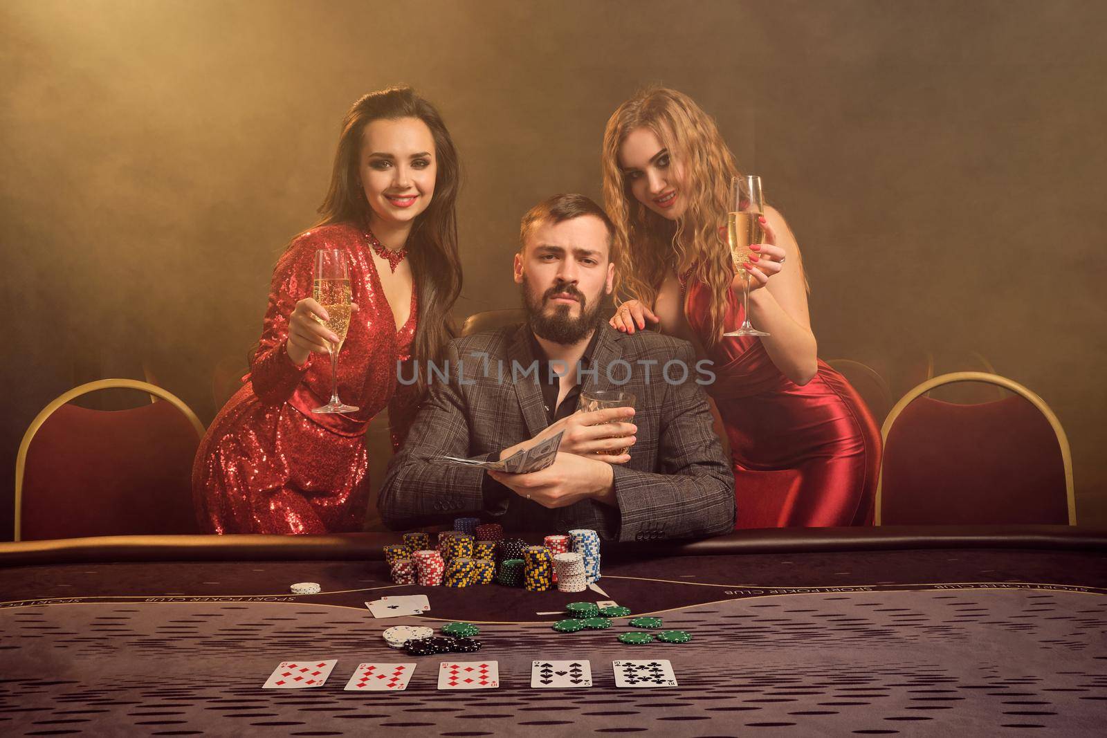 Two attractive girls and good-looking guy are playing poker at casino. They are holding their cash winning, smiling and looking at the camera while posing at the table against a yellow backlight on smoke background. Cards, chips, money, gambling, entertainment concept.