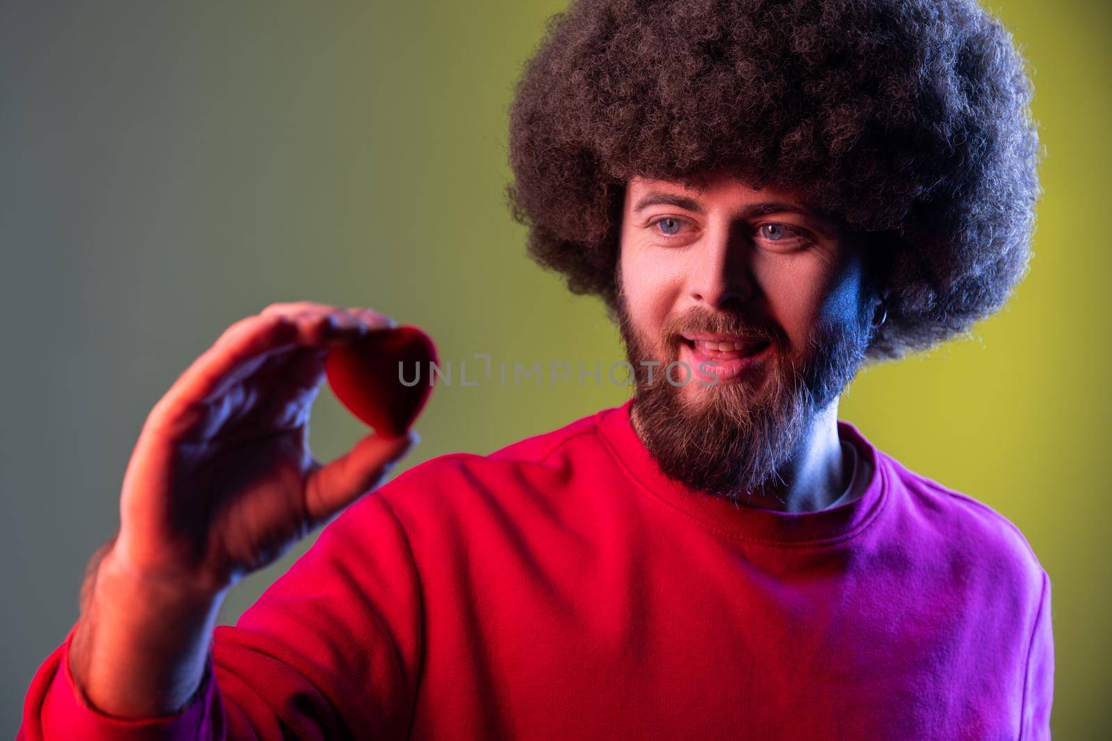 Portrait of hipster man with Afro hairstyle holding in hands little red heart confessing his love, romantic relationships. Indoor studio shot isolated on colorful neon light background.