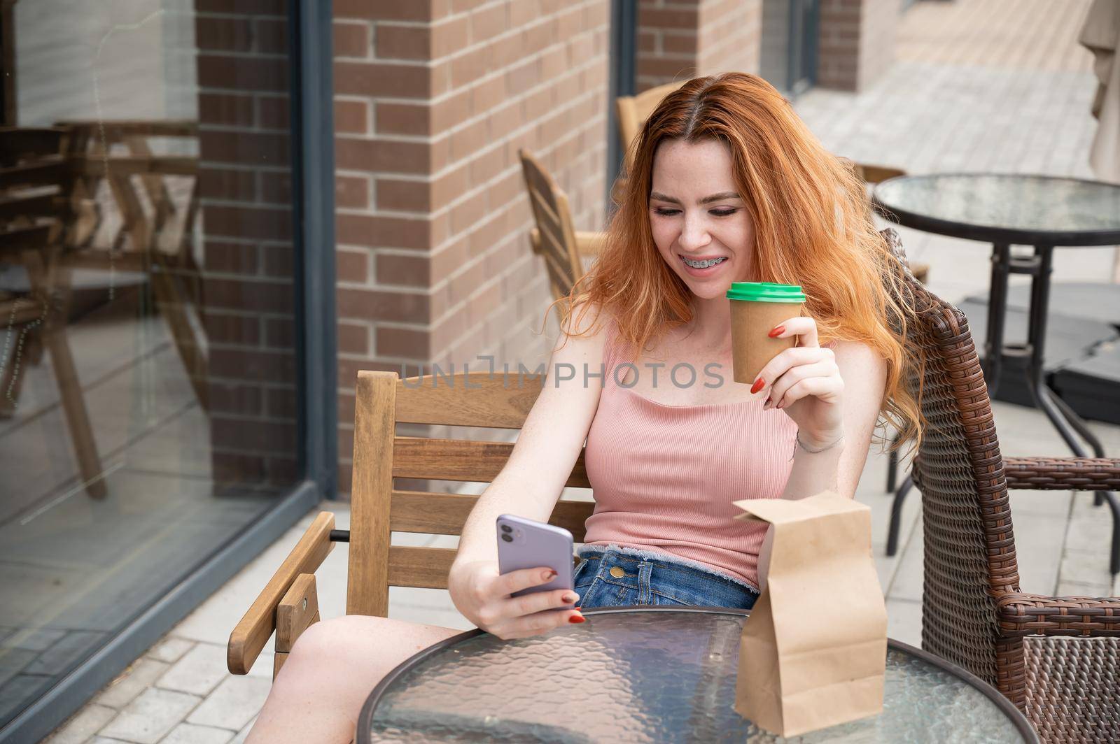 Young red-haired woman with braces on her teeth drinks coffee on a summer veranda