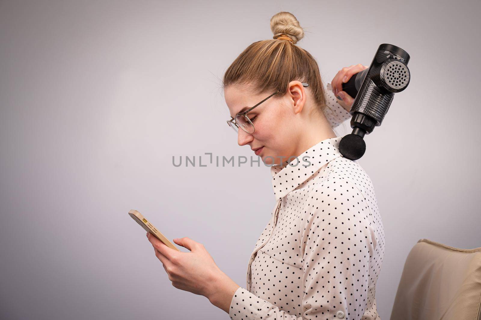 A young woman sits on a chair, uses a smartphone and makes herself massage with an electro massage gun