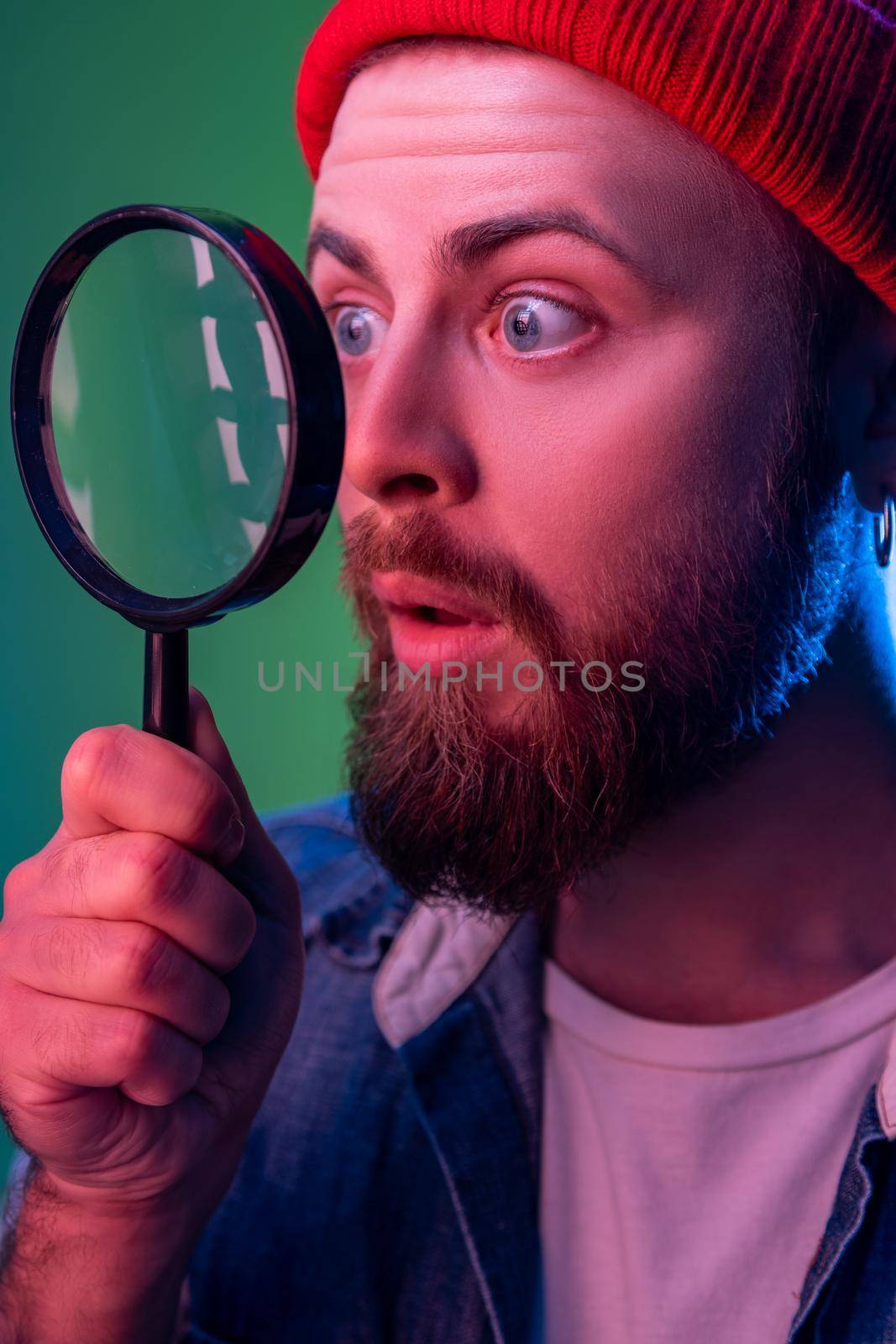 Side view of hipster man standing, holding magnifying glass and looking aside and having astonished shocked face. Indoor studio shot isolated on colorful neon light background.