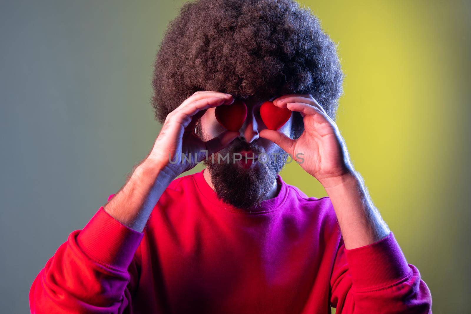 Portrait of hipster man with Afro hairstyle covering eyes with small red hearts, expressing romantic feelings, wearing red sweatshirt. Indoor studio shot isolated on colorful neon light background.