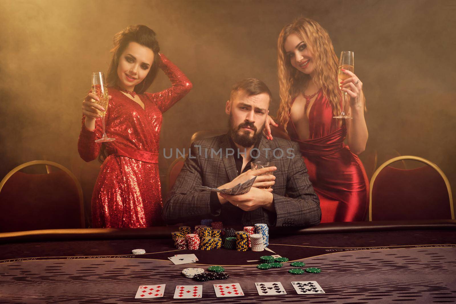Two attractive ladies and good-looking fellow are playing poker at casino. They are holding their cash winning, smiling and having a good time while posing at the table against a yellow backlight on smoke background. Cards, chips, money, gambling, entertainment concept.