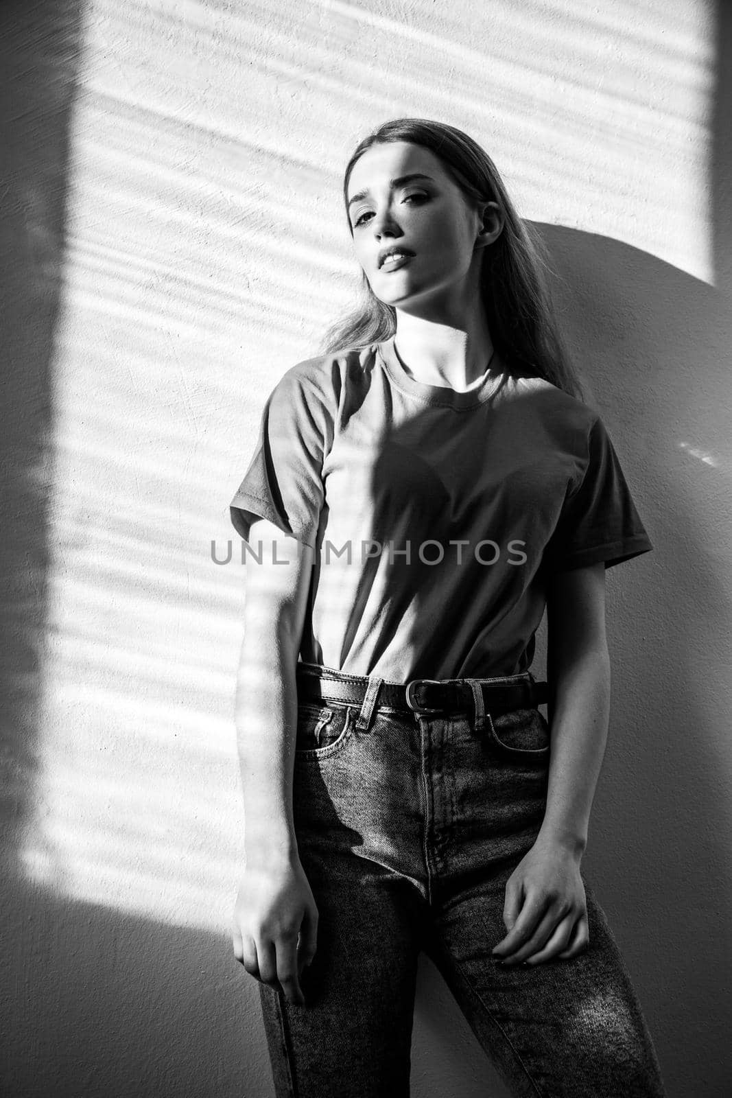 Young adult beautiful woman wearing T-shirt and jeans standing looking at camera with dreamy romantic expression. Black and white photography, indoor studio shot illuminated by sunlight from window