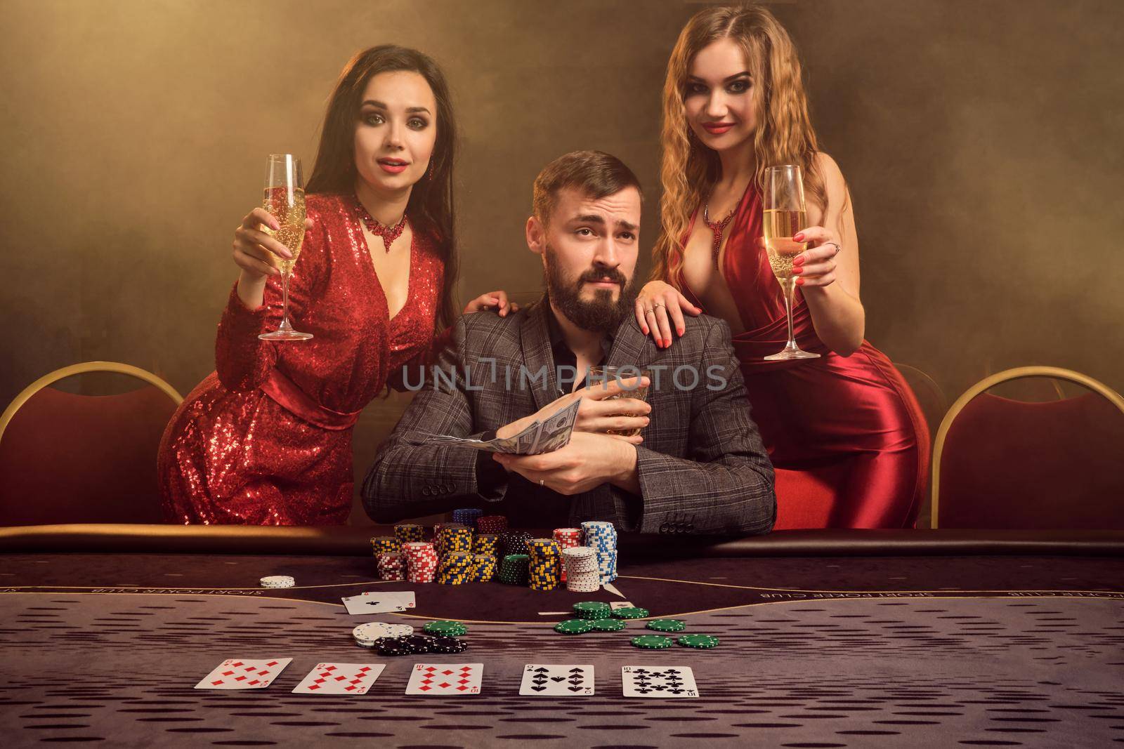 Two attractive maidens and good-looking man are playing poker at casino. They are holding their cash winning, smiling and looking happy while posing at the table against a yellow backlight on smoke background. Cards, chips, money, gambling, entertainment concept.