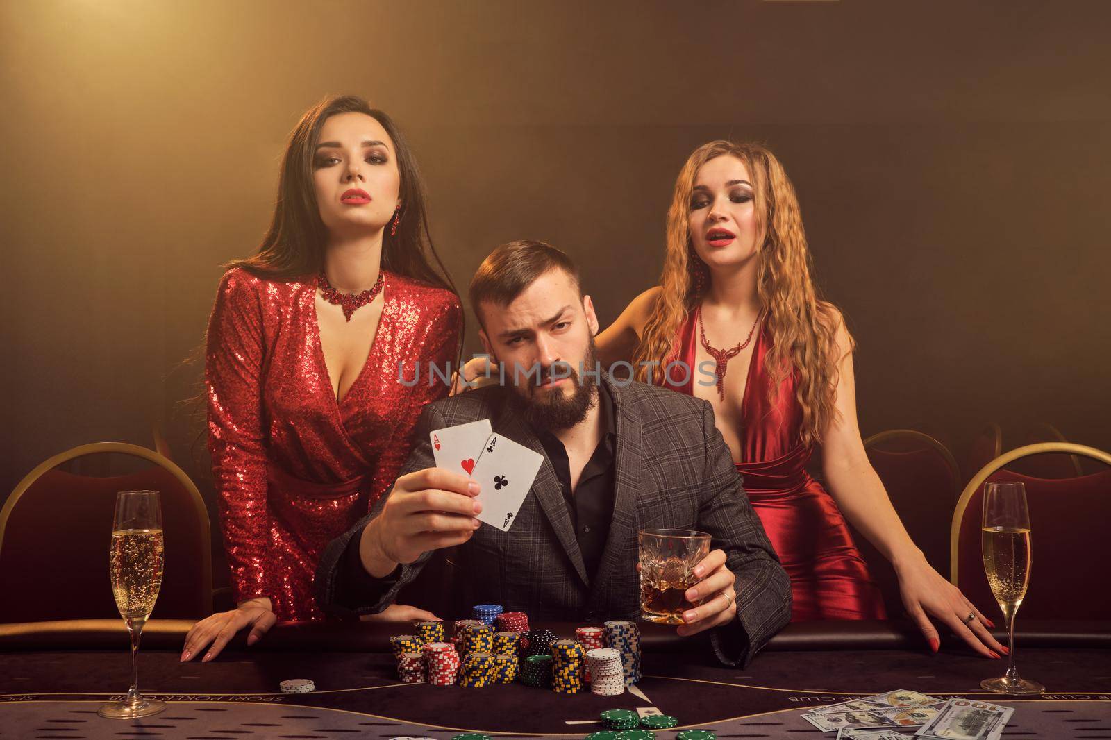 Two attractive women and good-looking man are playing poker at casino. They are showing the winning combination and looking at the camera while posing at the table against a yellow backlight on smoke background. Cards, chips, money, gambling, entertainment concept.