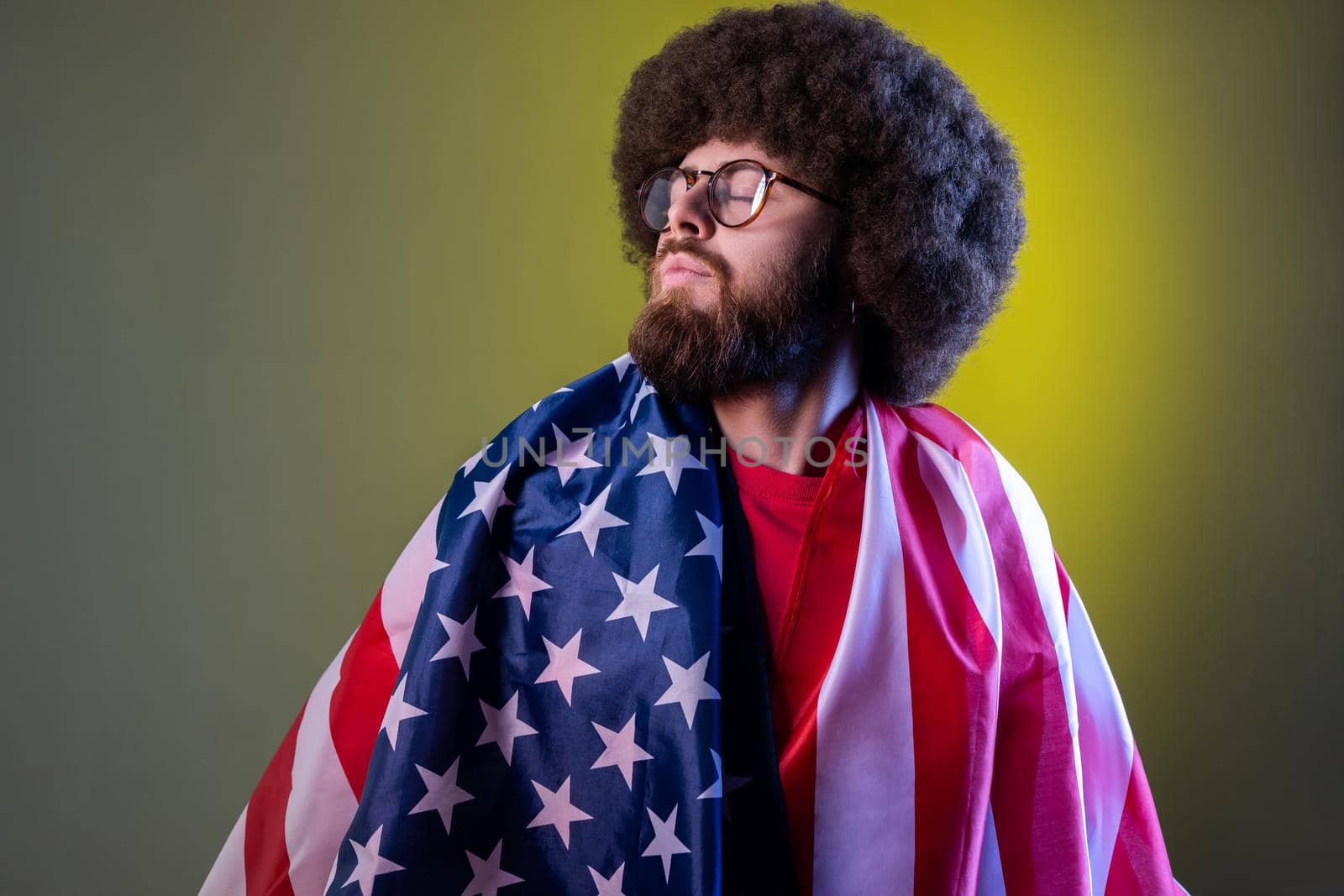 Portrait of proud hipster man with Afro hairstyle standing wrapped in american flag, looking away, feels pride of his USA. Indoor studio shot isolated on colorful neon light background.