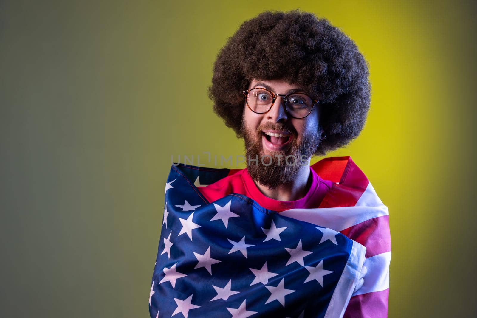 Portrait of hipster man standing wrapped in american flag, looking at camera with amazed excited facial expression. Indoor studio shot isolated on colorful neon light background.