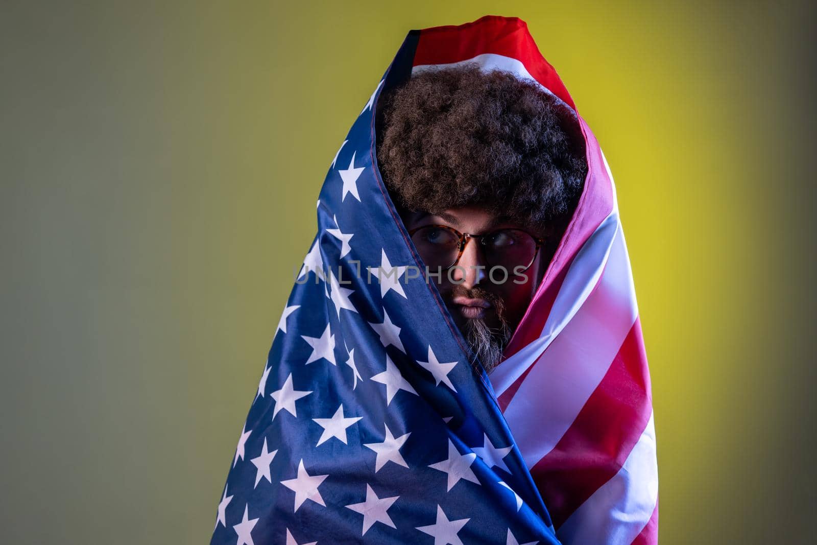 Portrait of hipster man with Afro hairstyle standing wrapped in american flag, looking away with funny facial expression and pout lips. Indoor studio shot isolated on colorful neon light background.