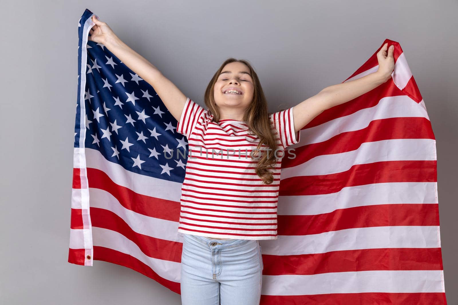 Portrait of extremely happy little girl wearing striped T-shirt holding USA flag over shoulders and keeps eyes closed and smiling happily. Indoor studio shot isolated on gray background.