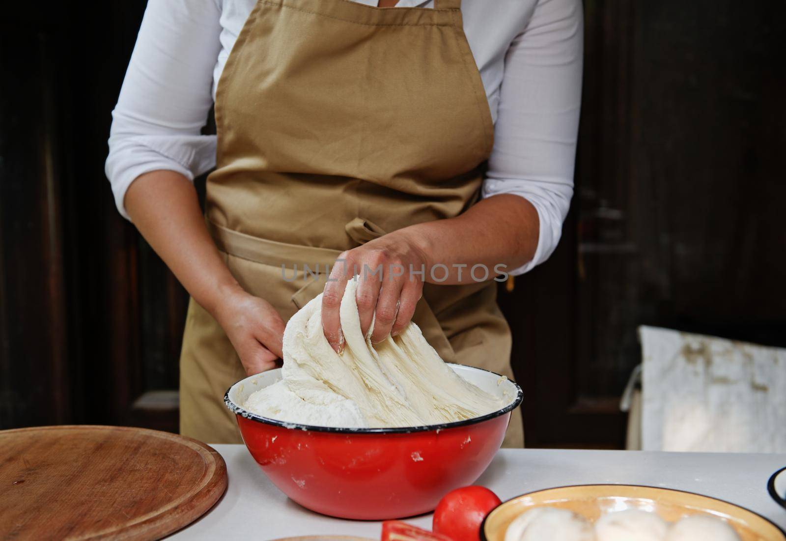 Details: Hands of chef in a beige apron kneading a raising yeast dough in a vintage enamel red bowl while preparing healthy whole grain bread of delicious Italian traditional pizza in a rustic kitchen