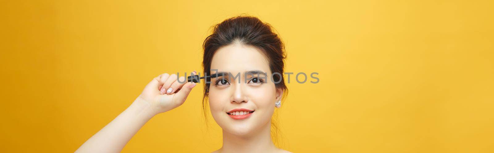 Closeup portrait of attractive young woman putting some mascara onto her eyelashes with make up brush over yellow background by makidotvn