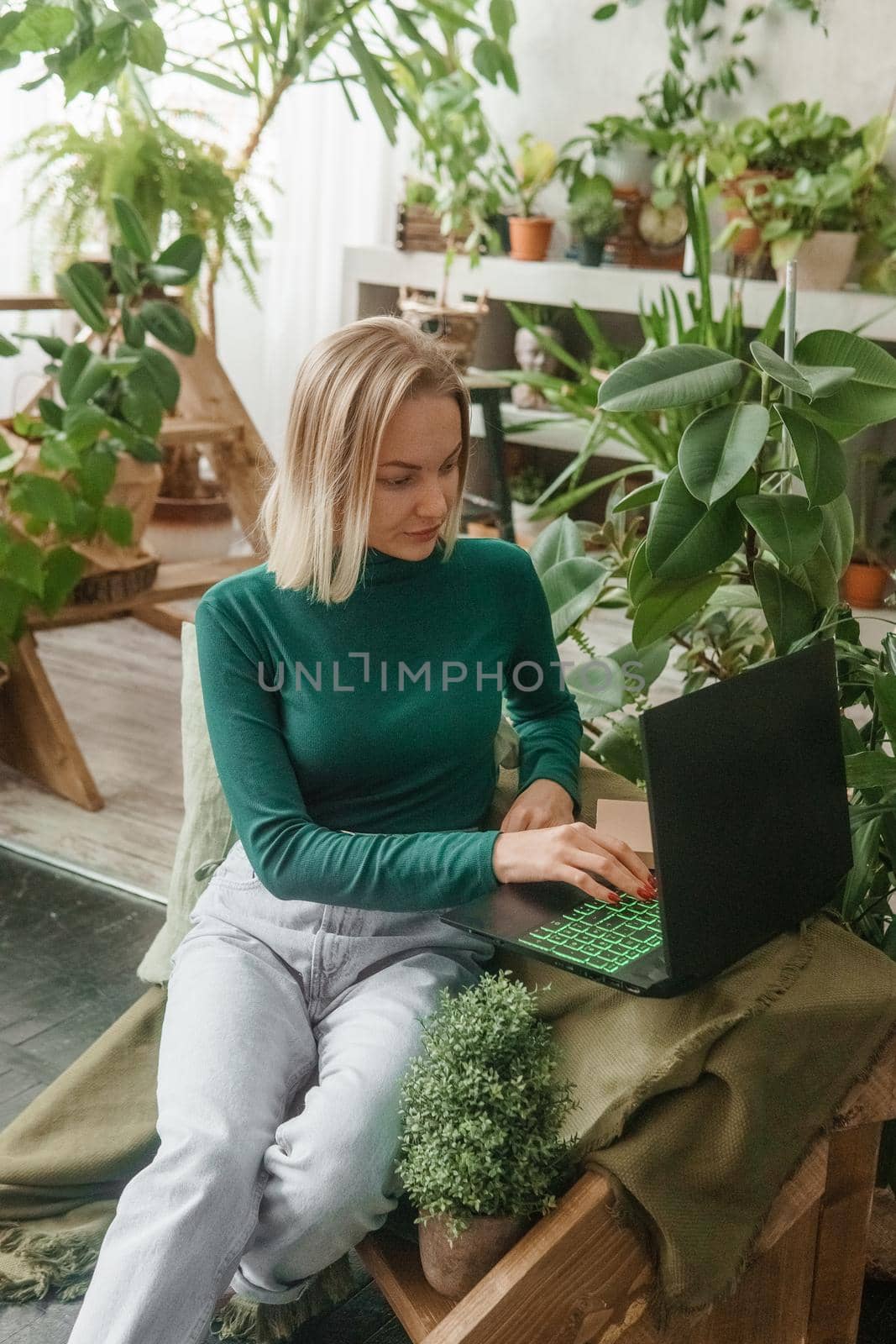 A blonde woman in a room with a lot of green indoor plants is working on a laptop. The concept of biophysical design in the interior. Work from home, work as a freelancer.