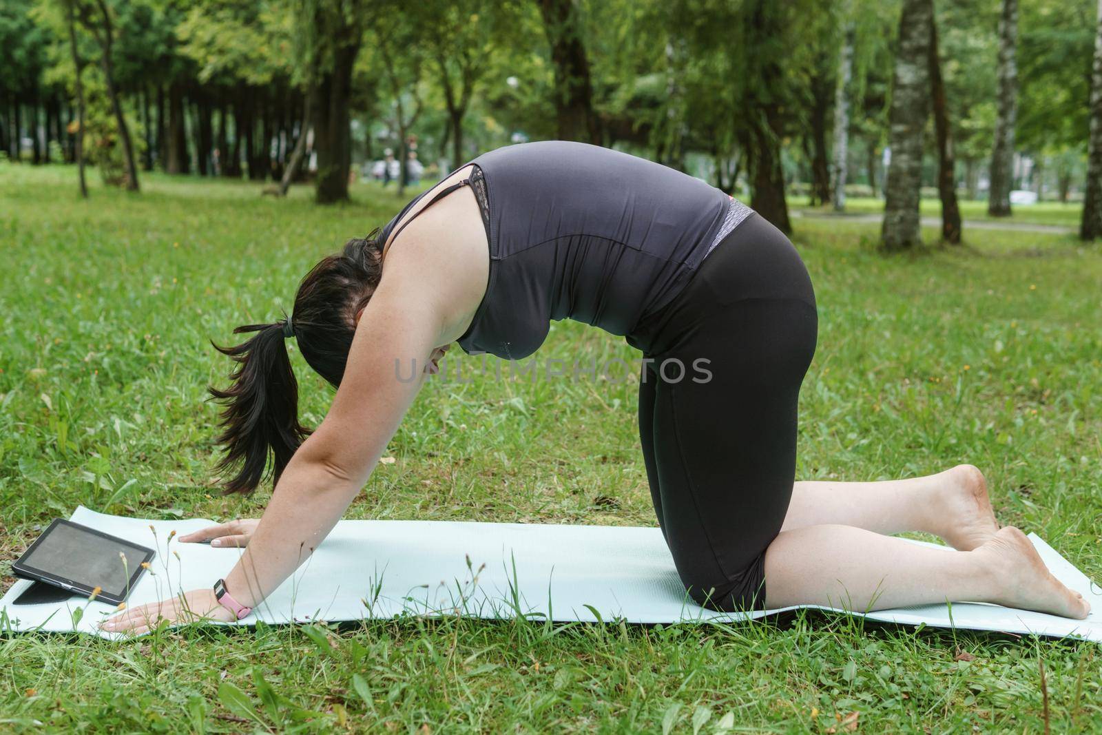 A charming brunette woman plus-size body positive practices sports in nature. Woman does yoga in the park on a sports mat. On a yoga mat in the dog pose.