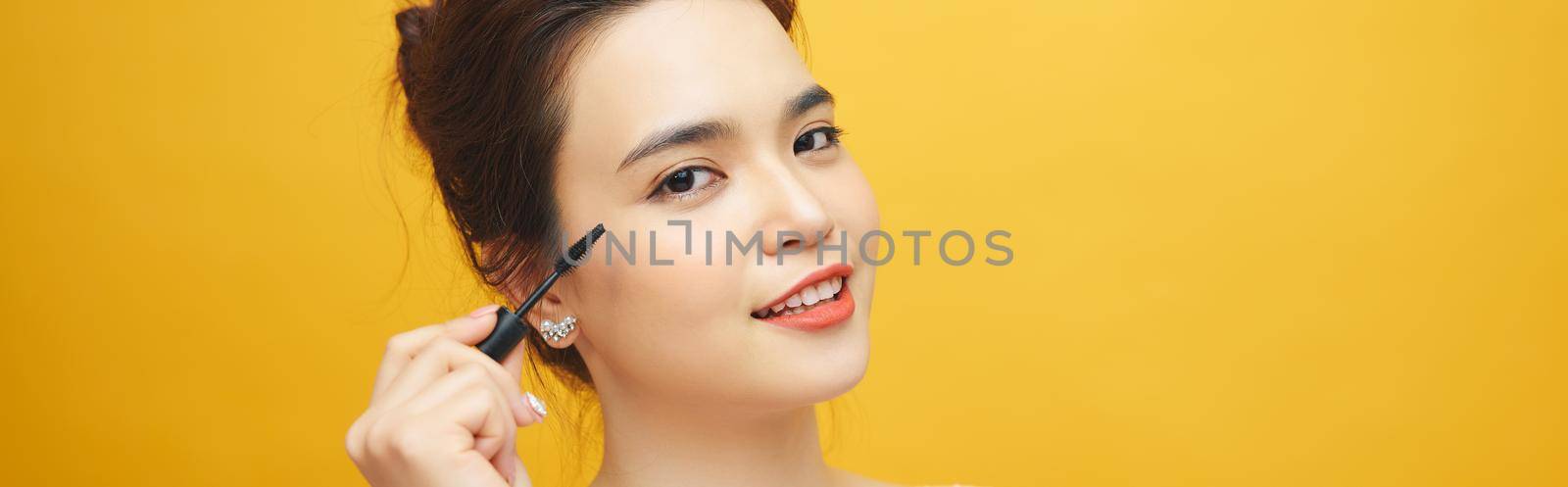 Closeup portrait of attractive young woman putting some mascara onto her eyelashes with make up brush over yellow background