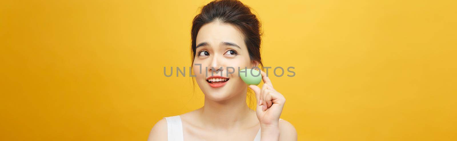 Beautiful young woman applying makeup using beauty blender sponge. Isolated over yellow background  by makidotvn