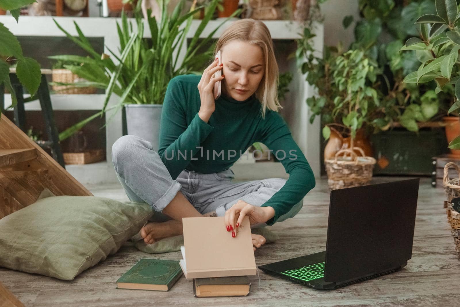 A blonde woman in a room with a lot of green indoor plants is working on a laptop. The concept of biophysical design in the interior. Work from home, work as a freelancer.