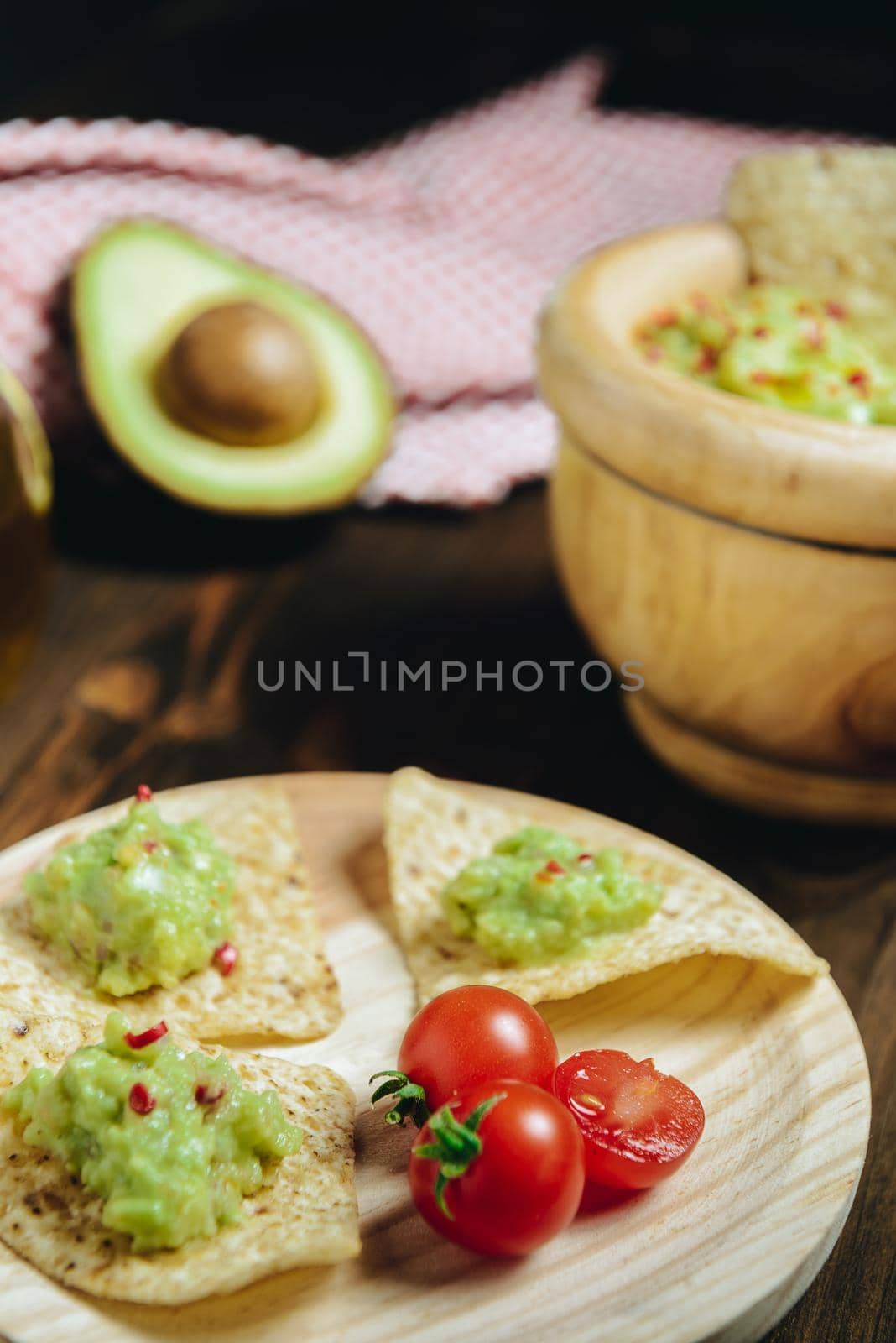 vertical photo of fresh guacamole in a wood bowl with nachos next to an avocado and a kitchen rag, typical mexican healthy vegan cuisine with rustic dark food photo style
