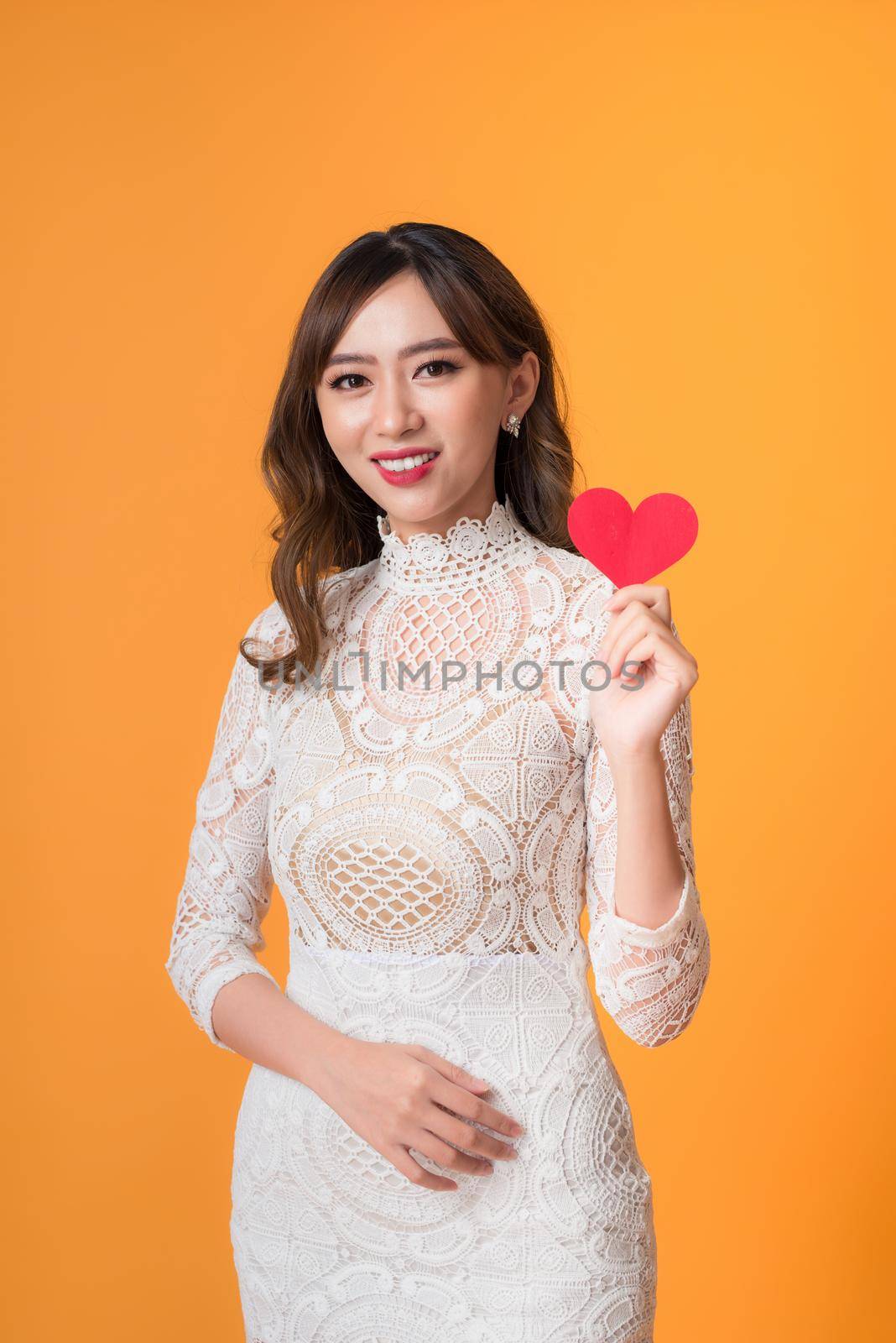 Portrait of young beautiful asian woman hands holding red heart shape isolated on yellow background.