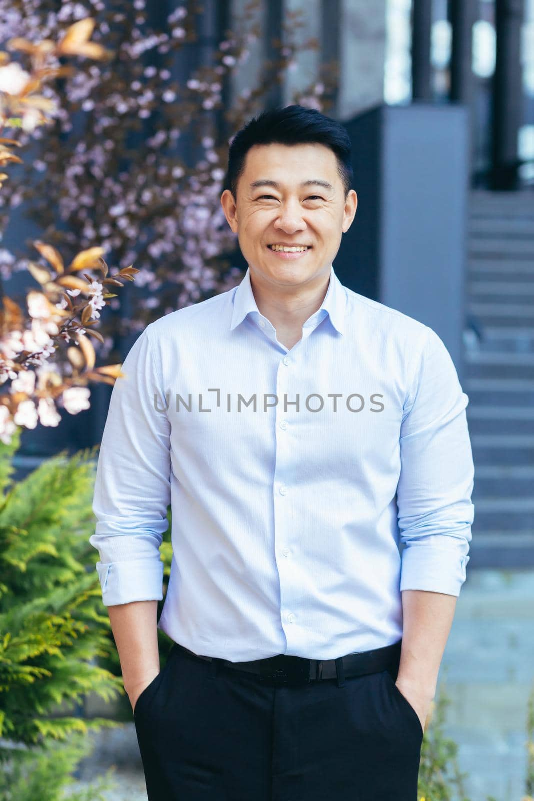 Vertical frame, Asian man in white shirt looks at camera and smiles, hands in pockets