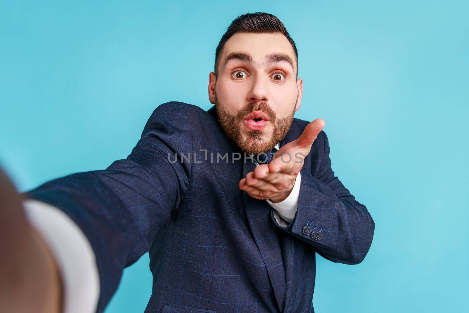 Handsome young adult man wearing official style suit taking selfie, looking at camera POV, point of view of photo, sending air kiss. Indoor studio shot isolated on blue background.