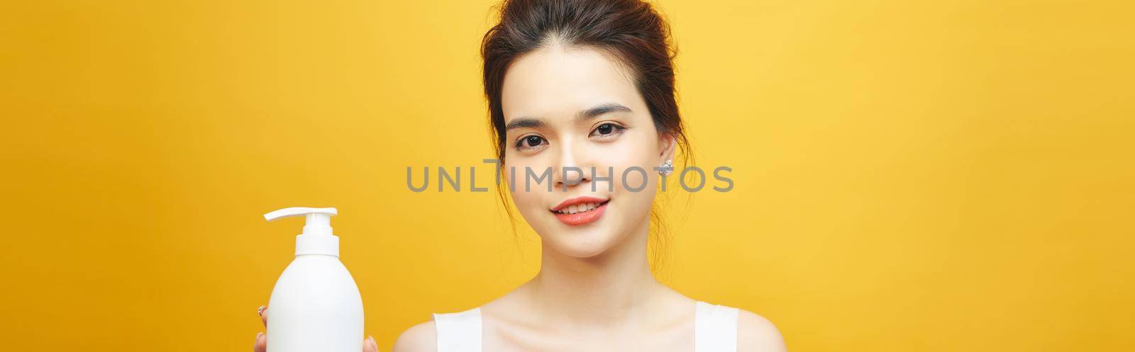 Smiling young asian woman showing skincare product on yellow background