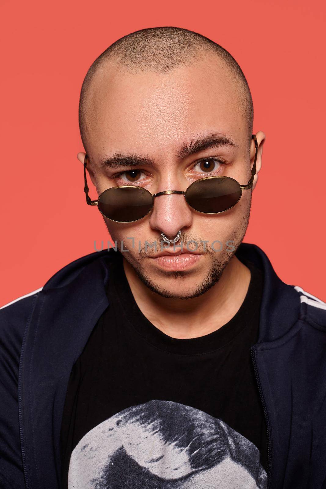 Close-up studio shot of a handsome tattoed bald person in sunglasses, with a pirsing ring in his nose, wearing black trendy t-shirt with print and sport suit, looking at you while posing against a pink background with copy space. People, style and fashion concept. 90s style