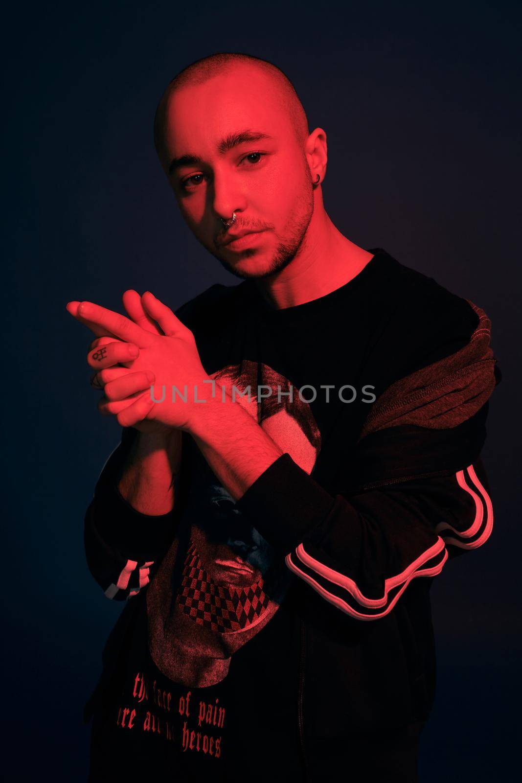 Close-up studio shot of a good-looking tattoed bald fellow with a pirsing ring in his nose, wearing black trendy t-shirt with print and sport suit, folded his arms like a gun posing against a dark blue background with copy space. People, style and fashion concept. 90s style