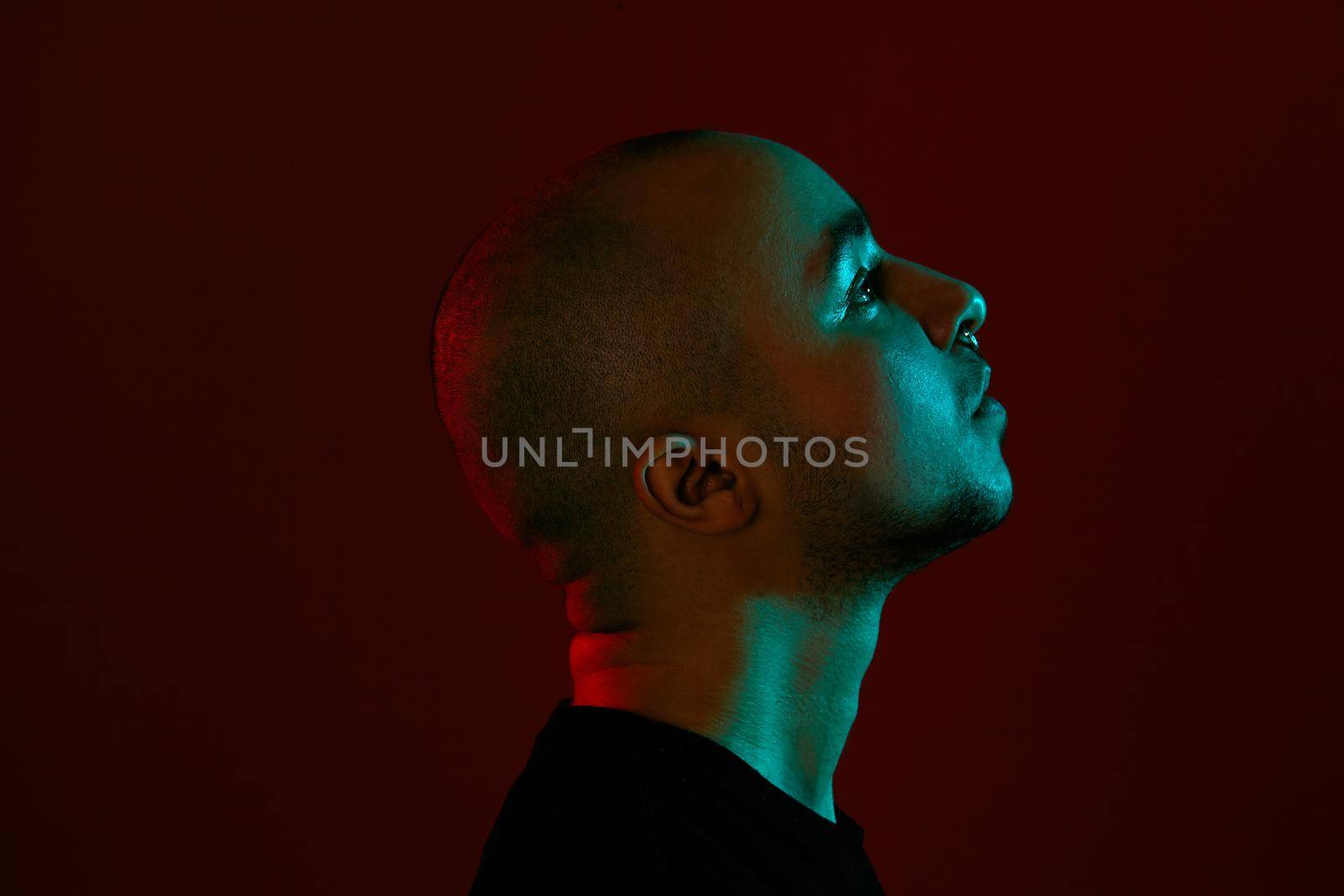 Close-up studio shot of a handsome tattoed bald male with a pirsing ring in his nose, wearing black trendy t-shirt with print, posing sideways against a red background with copy space. People, style and fashion concept. 90s style