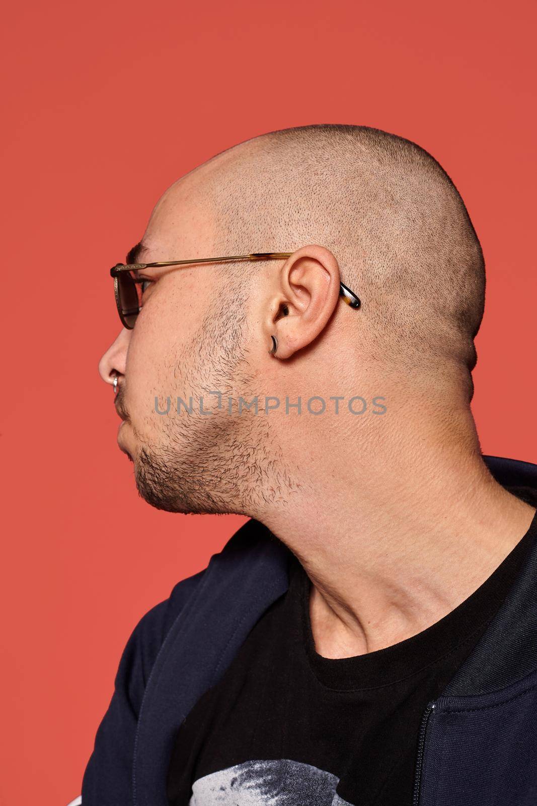 Close-up studio shot of a relaxed tattoed bald guy in sunglasses, with a pirsing ring in his nose, wearing black trendy t-shirt with print and sport suit, posing sideways against a pink background with copy space. People, style and fashion concept. 90s style