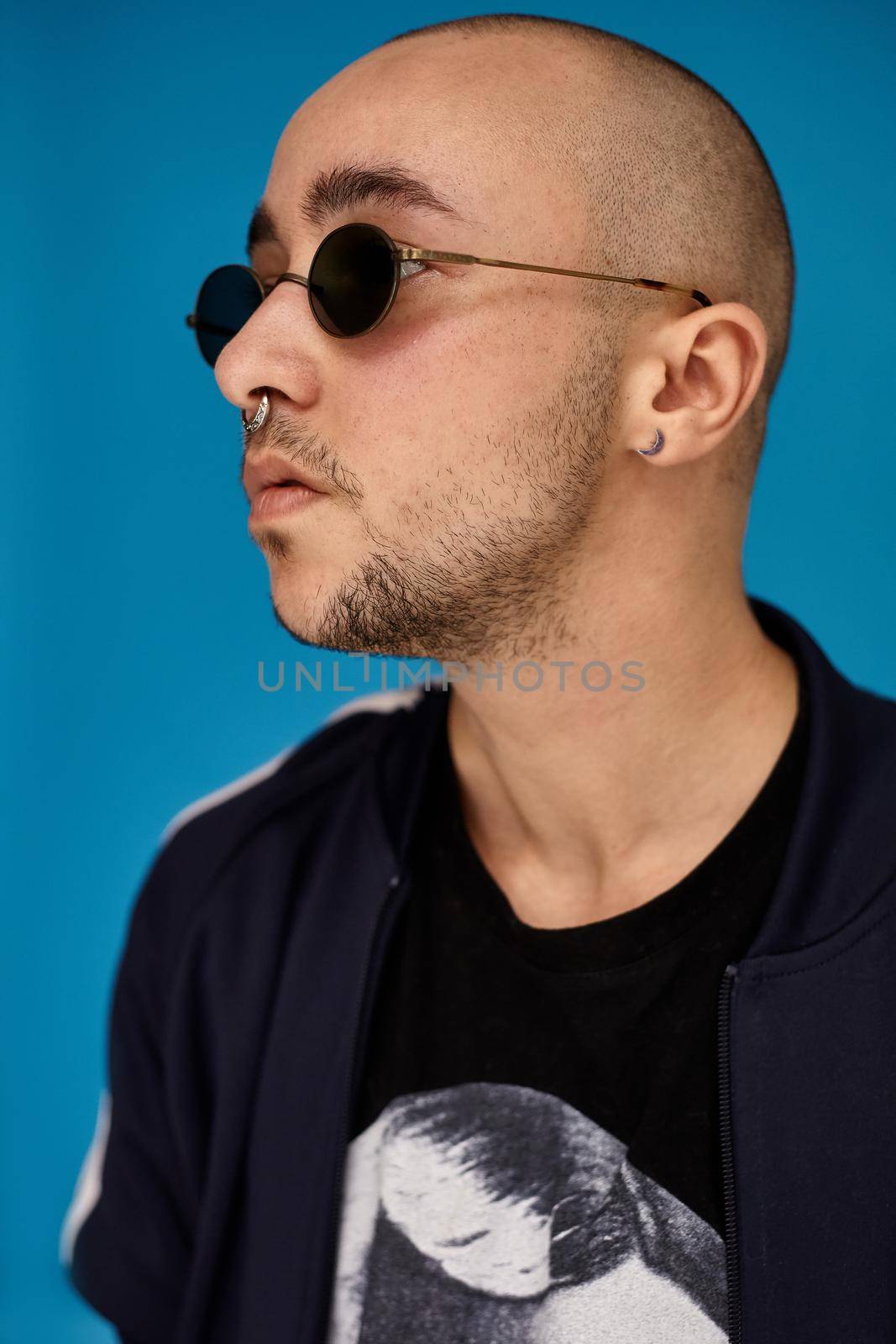 Close-up studio shot of a young tattoed bald guy in sunglasses, with a pirsing ring in his nose, wearing black trendy t-shirt with print and sport suit, posing sideways against a blue background with copy space. People, style and fashion concept. 90s style