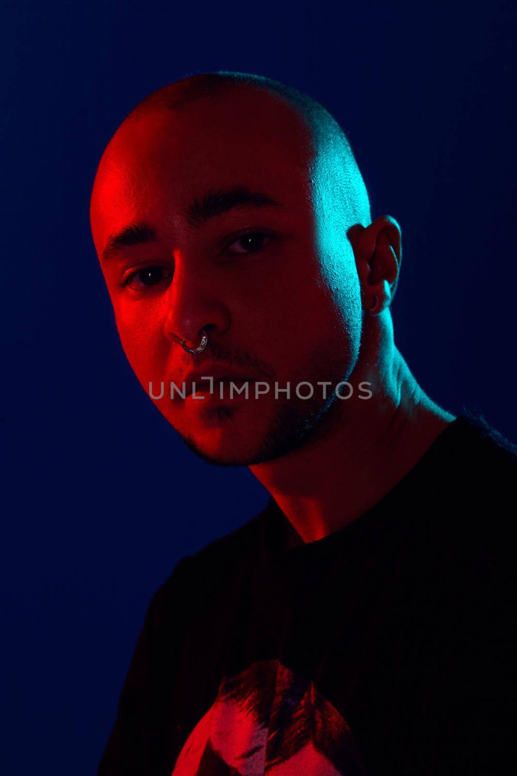 Close-up studio shot of a good-looking tattoed bald person with a pirsing ring in his nose, wearing black trendy t-shirt with print, posing and looking at the camera against a blue background with copy space. People, style and fashion concept. 90s style