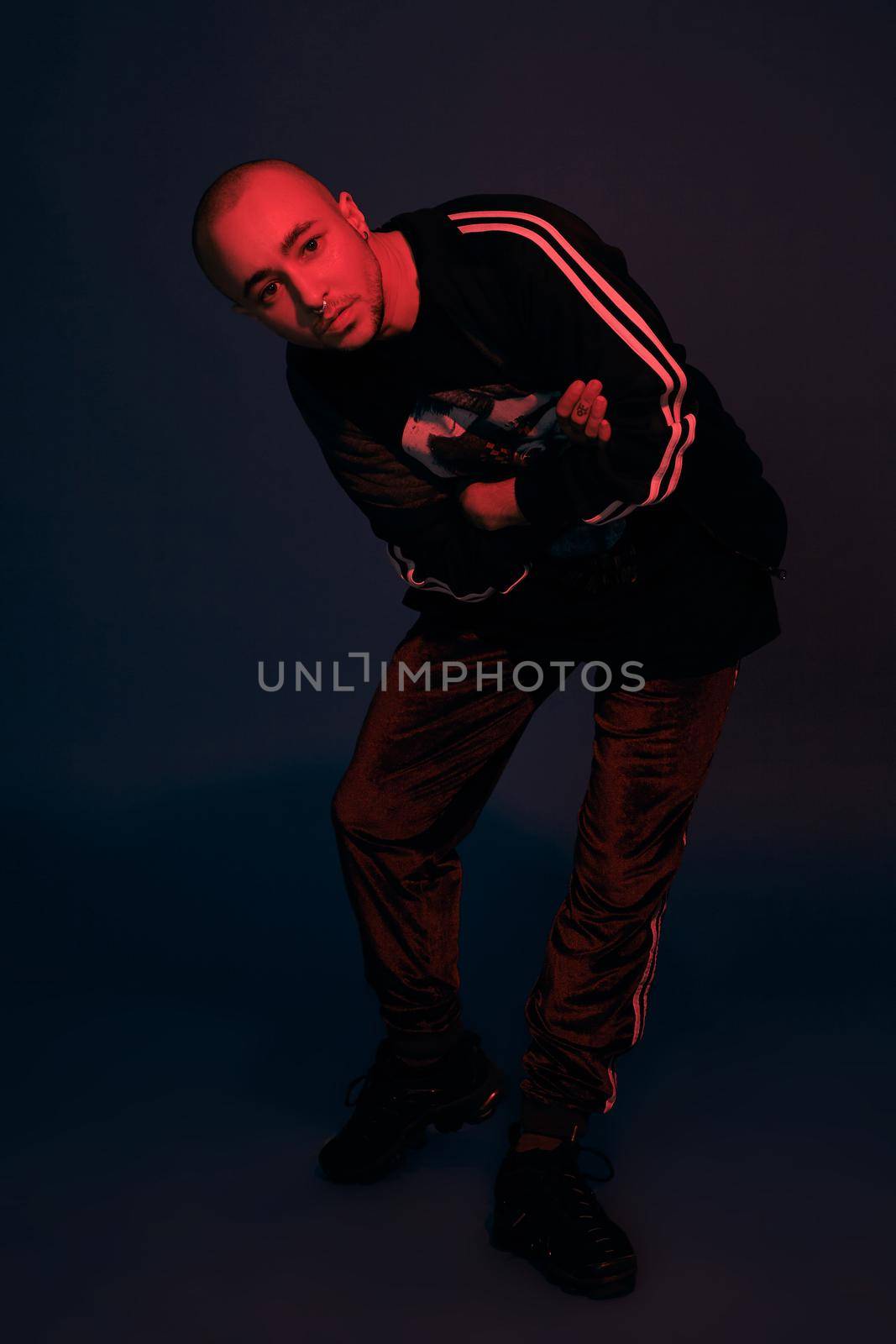 Full-length studio shot of a handsome tattoed bald person with a pirsing ring in his nose, wearing black trendy t-shirt with print, sport suit and sneakers, looking at the camera while posing with crossed hands against a dark blue background with copy space. People, style and fashion concept. 90s style