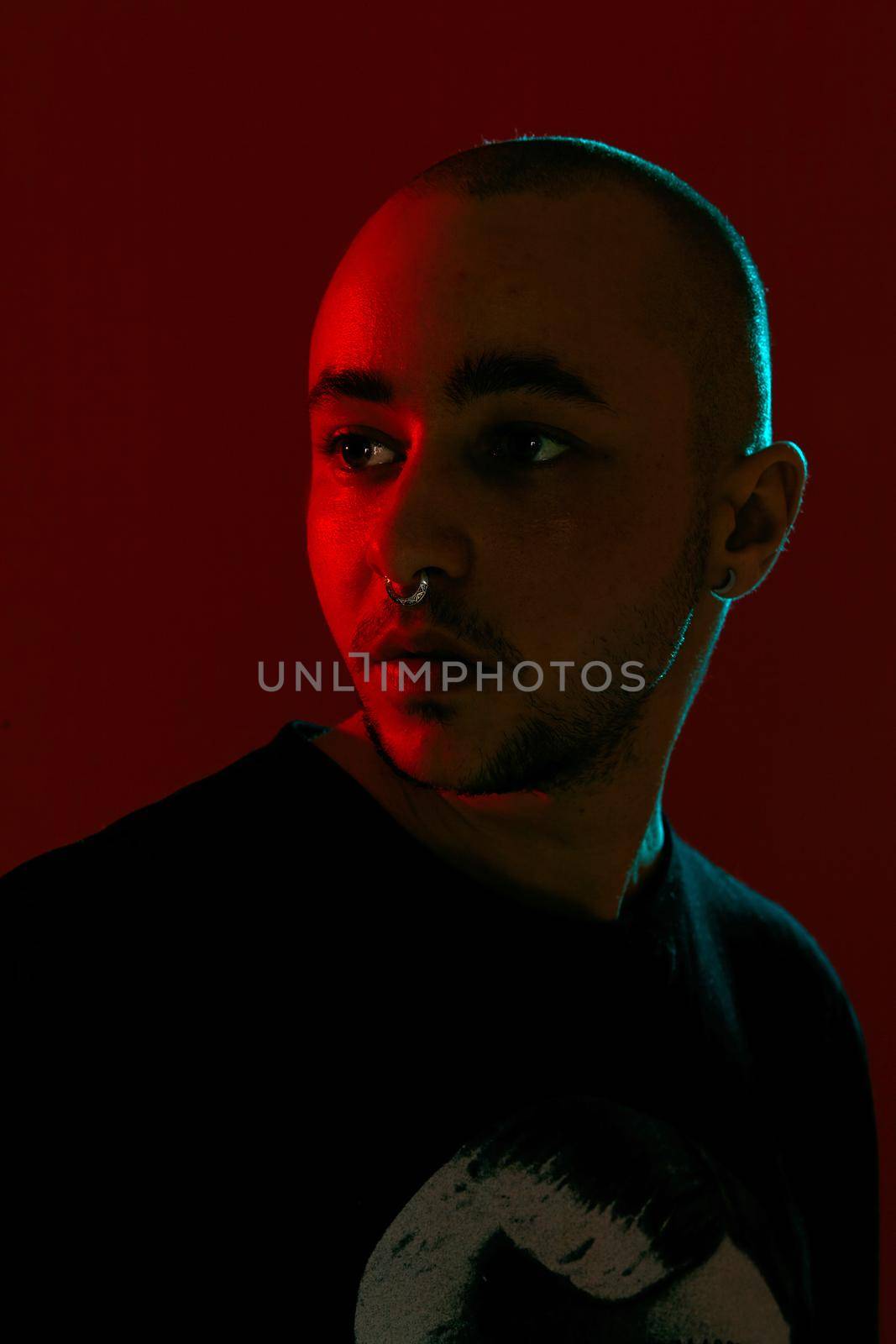 Close-up studio shot of a good-looking tattoed bald male with a pirsing ring in his nose, wearing black trendy t-shirt with print, posing against a red background with copy space. People, style and fashion concept. 90s style