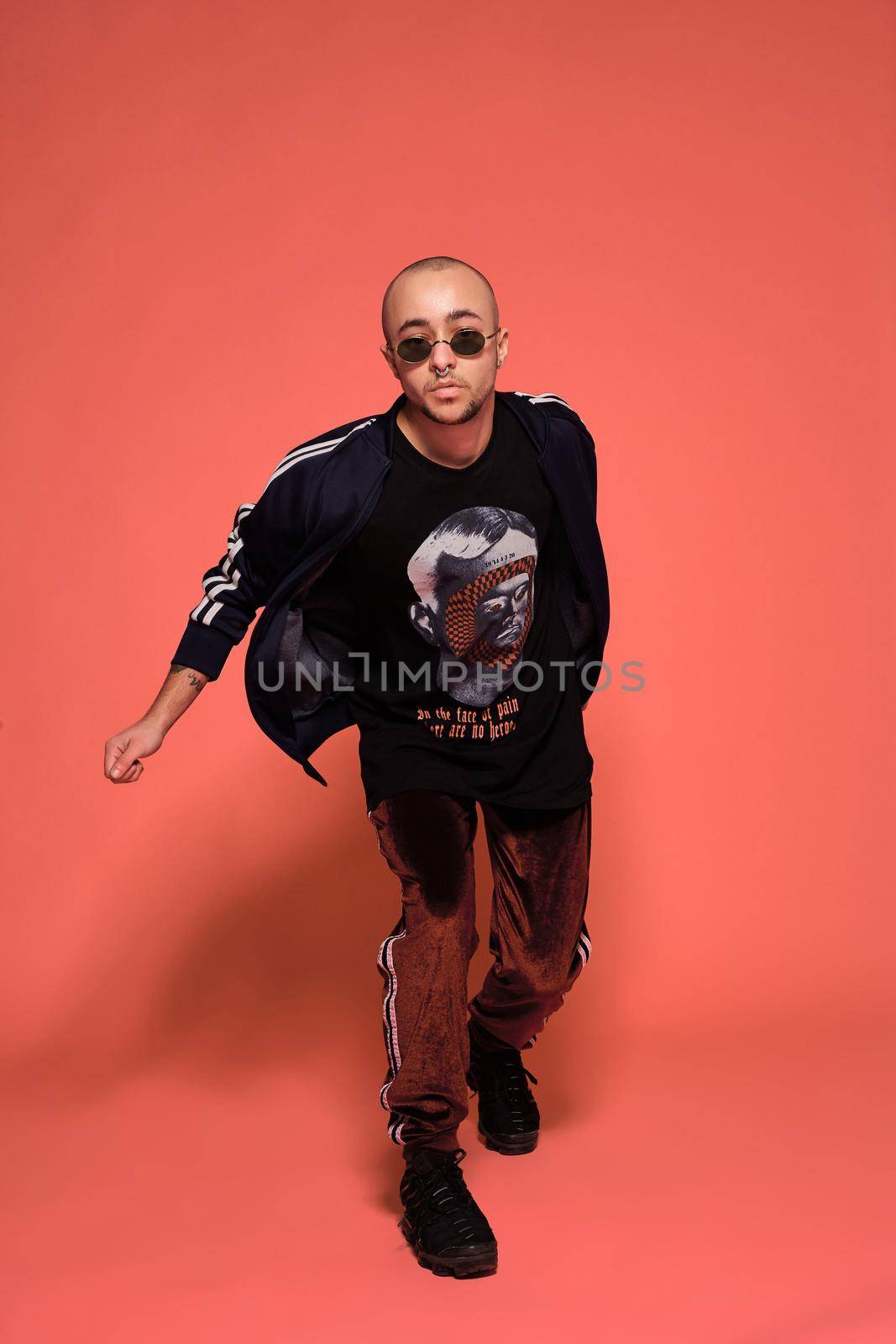 Full-length studio shot of a young tattoed bald person in sunglasses, with a pirsing ring in his nose, wearing black trendy t-shirt with print, sport suit and sneakers posing against a pink background with copy space. People, style and fashion concept. 90s style