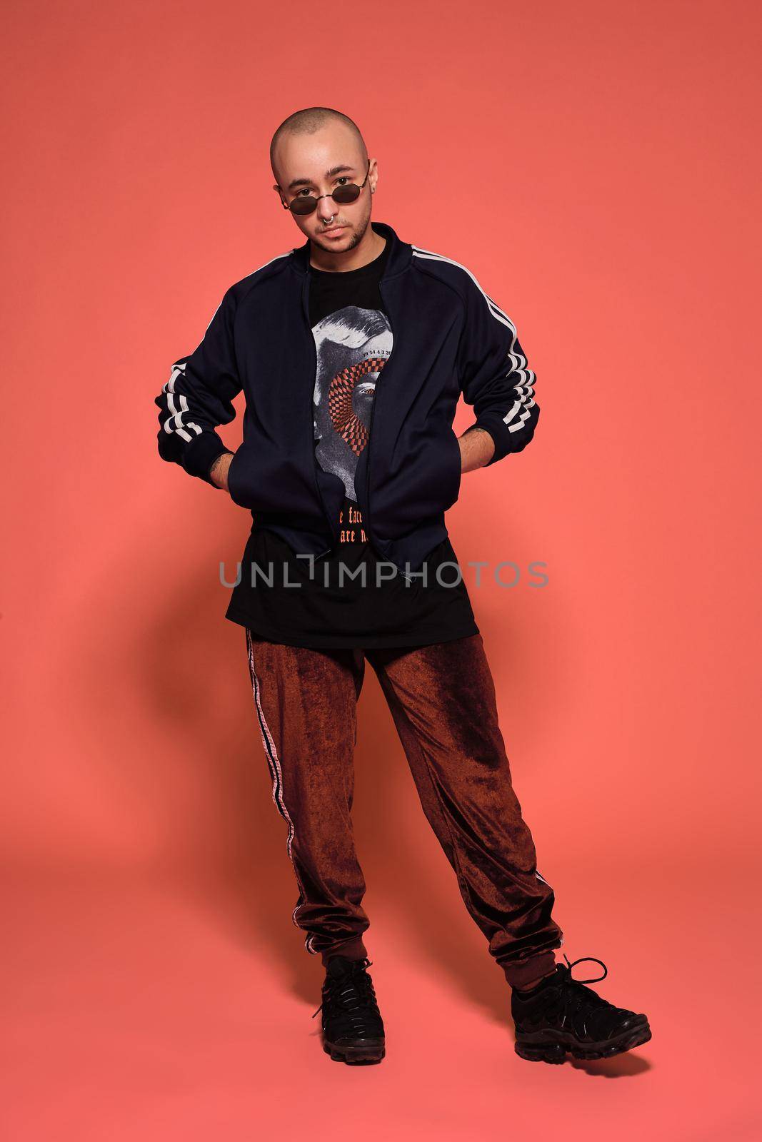 Full-length studio shot of a young tattoed bald guy in sunglasses, with a pirsing ring in his nose, wearing black trendy t-shirt with print, sport suit and sneakers posing against a pink background with copy space. People, style and fashion concept. 90s style