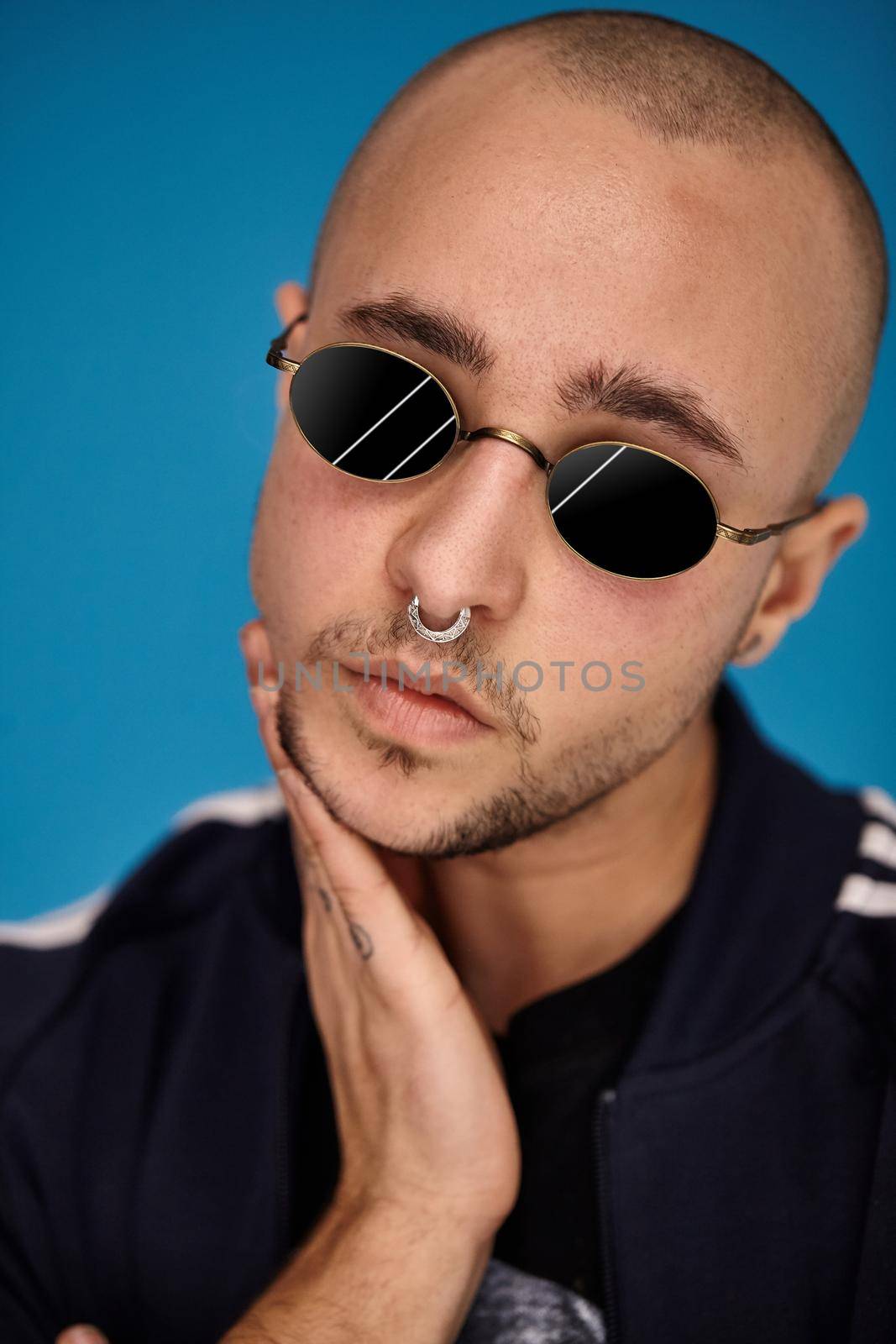 Close-up studio shot of a good-looking tattoed bald person in sunglasses, with a pirsing ring in his nose, wearing black trendy t-shirt with print and sport suit, looking at the camera and posing against a blue background with copy space. People, style and fashion concept. 90s style