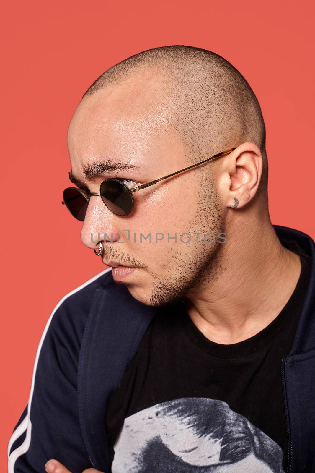 Close-up studio shot of a relaxed tattoed bald fellow in sunglasses, with a pirsing ring in his nose, wearing black trendy t-shirt with print and sport suit, looking away and posing against a pink background with copy space. People, style and fashion concept. 90s style