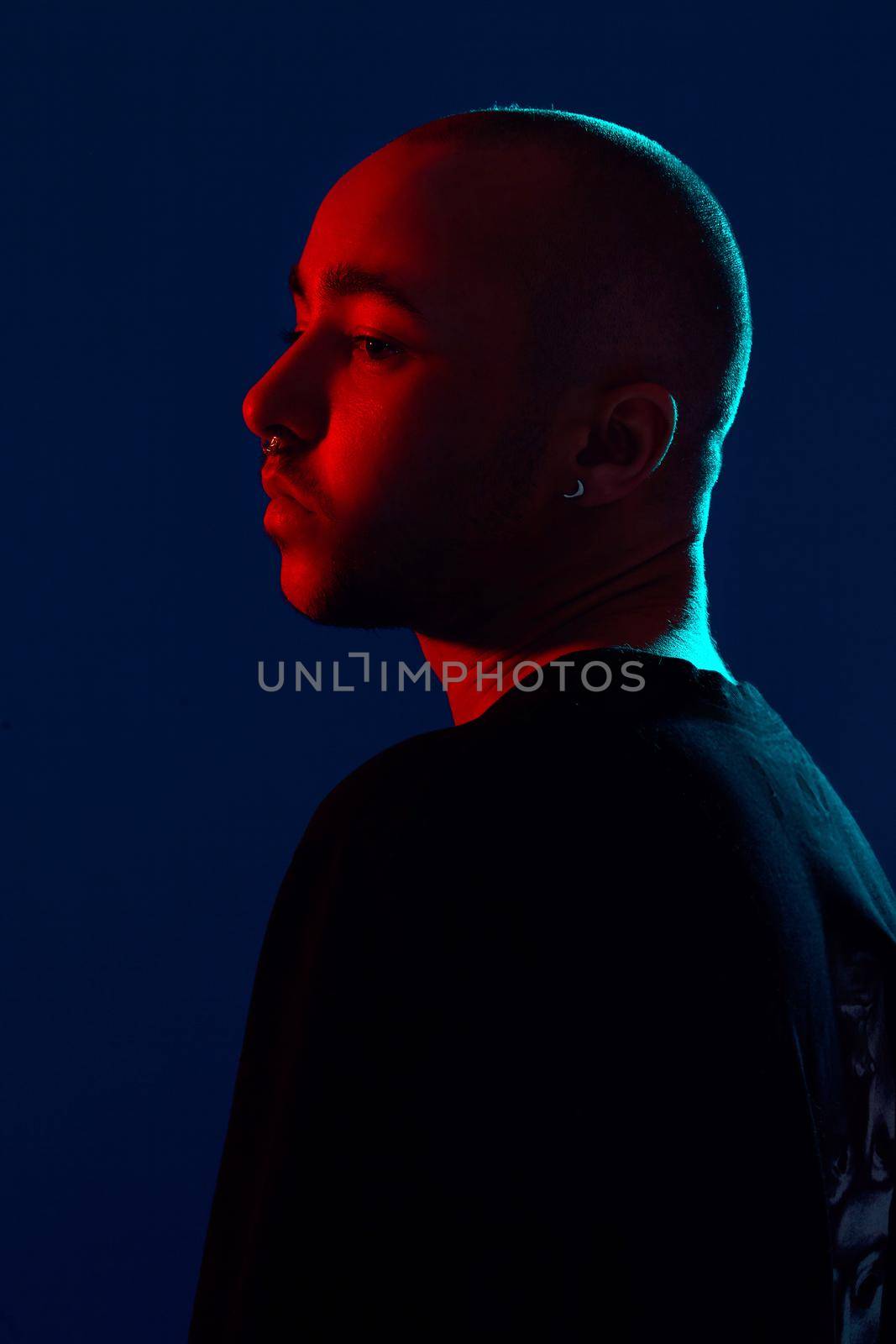 Close-up studio shot of a relaxed tattoed bald male with a pirsing ring in his nose, wearing black trendy t-shirt with print, posing sideways against a blue background with copy space. People, style and fashion concept. 90s style