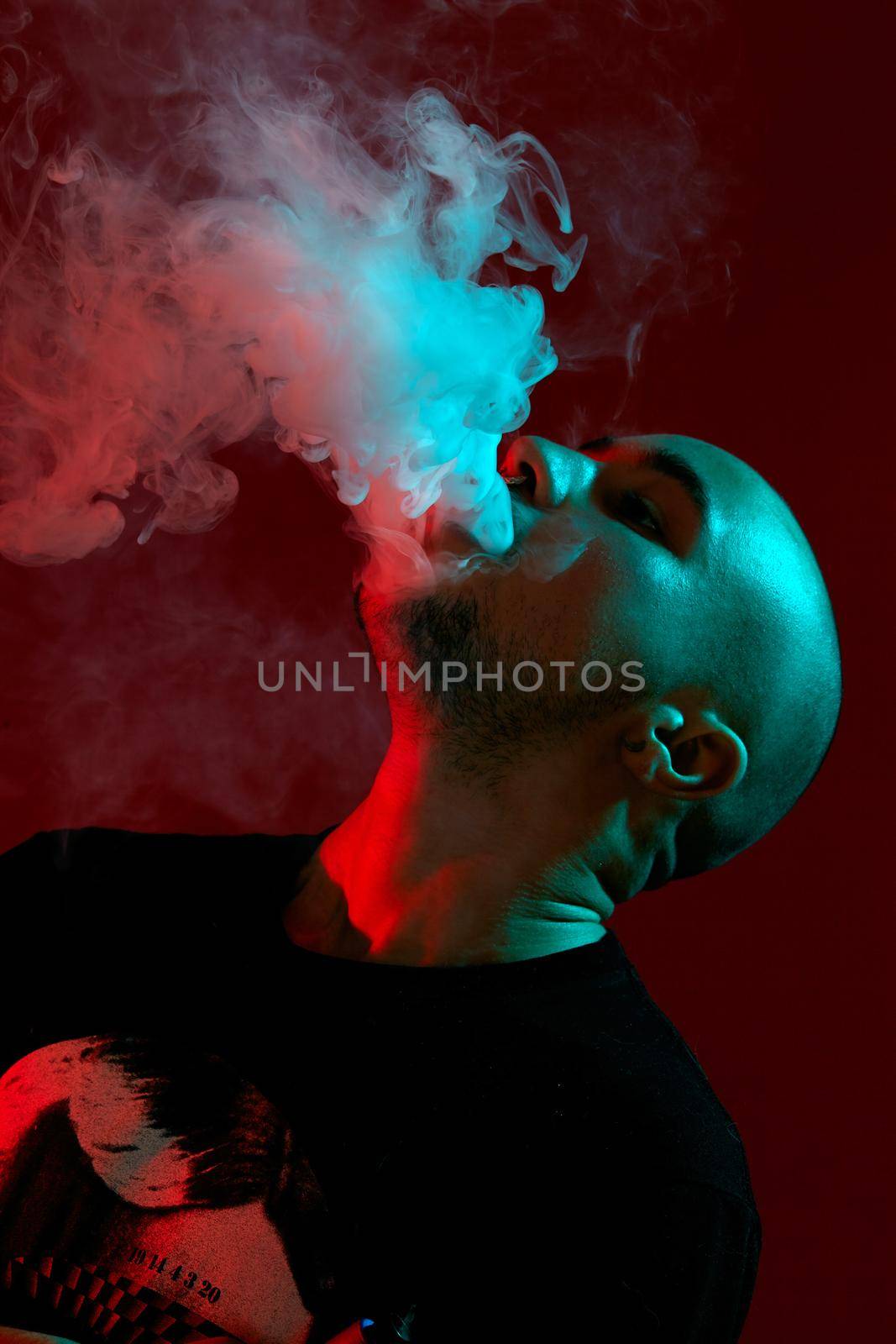 Close-up studio shot of a handsome tattoed bald person with a pirsing ring in his nose, in a black t-shirt with print, thoughtfully and calmly vaping, blowing out a cloud of smoke on burgundy background with backlight. Vape addiction concept, nicotine free smoking, copy space. 90s style