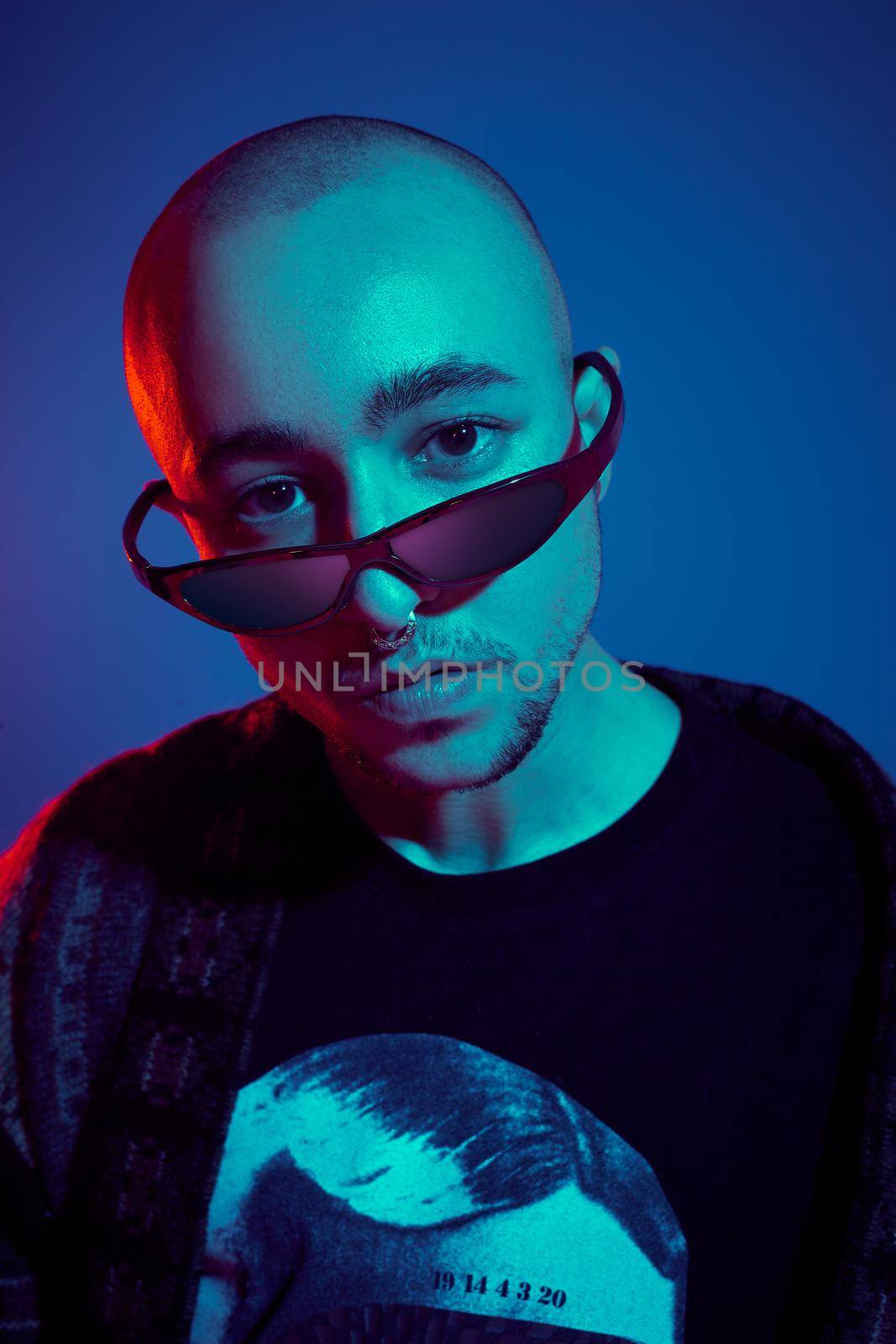 Close-up studio shot of a enjoying tattoed bald fellow in sunglasses, with a pirsing ring in his nose, wearing dark jacket, black trendy t-shirt with print, looking at the camera and posing against a blue background with copy space. People, style and fashion concept. 90s style