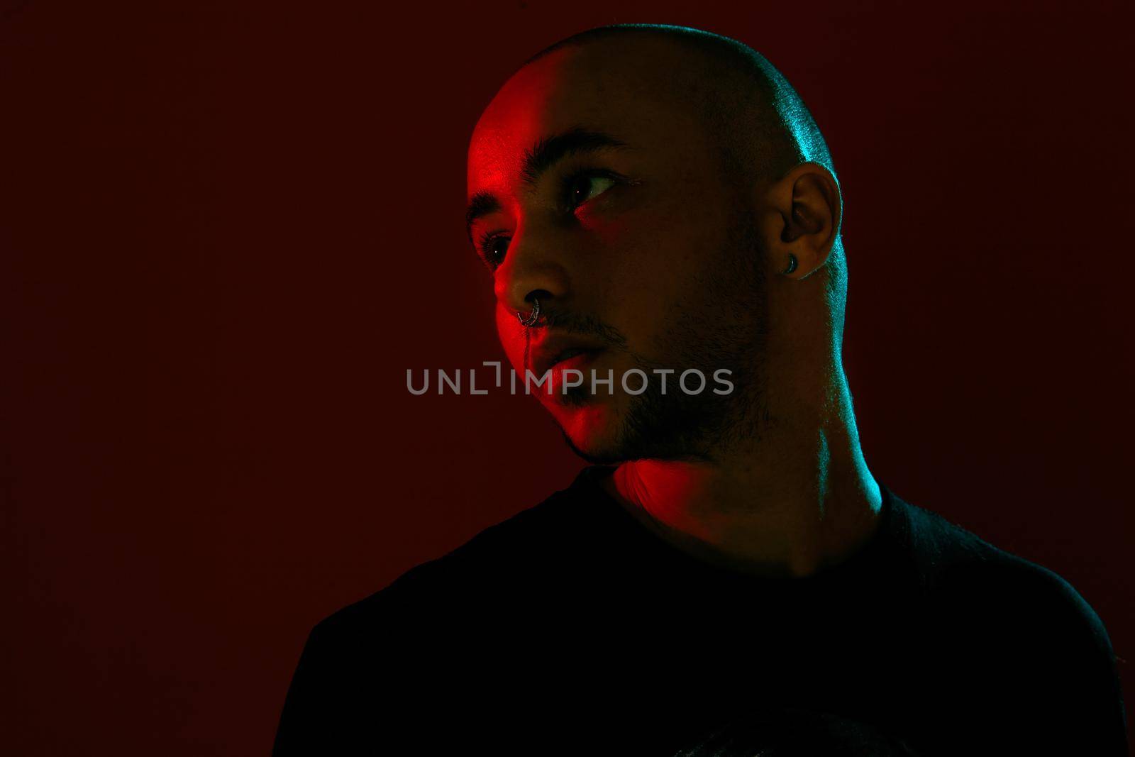 Close-up studio shot of a good-looking tattoed bald guy with a pirsing ring in his nose, wearing black trendy t-shirt with print, posing and looking up against a red background with copy space. People, style and fashion concept. 90s style